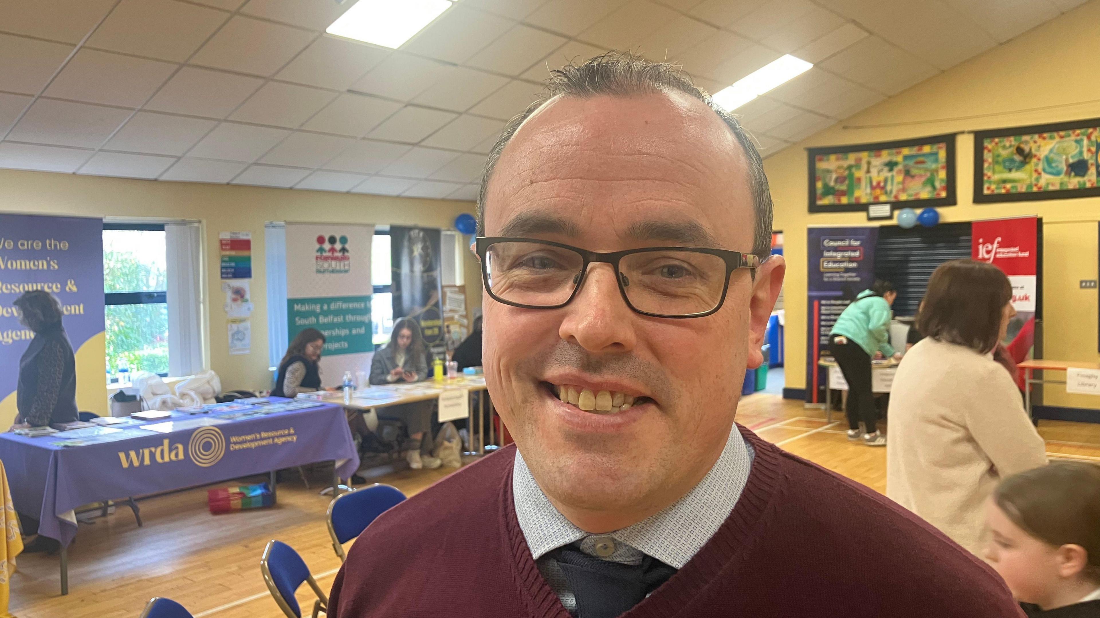 A man wearing reading glasses, a blue shirt, navy tie and burgundy sweater is smiling at the camera. Behind him are information stalls set up in a school hall