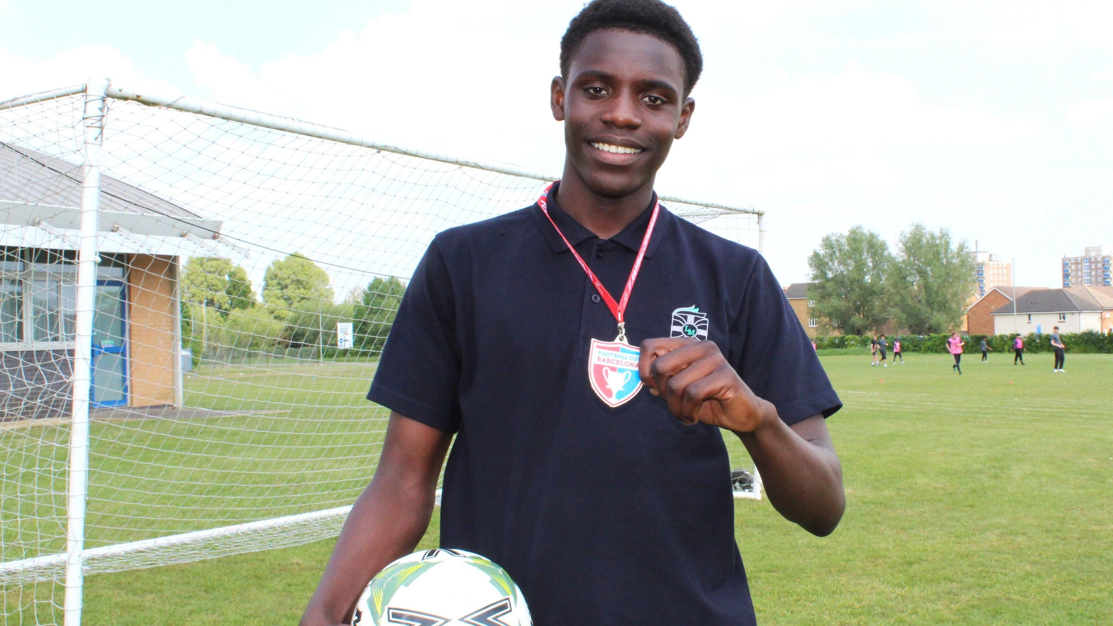 Bubacarr smiles at the camera while standing on a football pitch. He has a polo top on and holds a football in one arm and a Barcelona badge and lanyard in the other.
