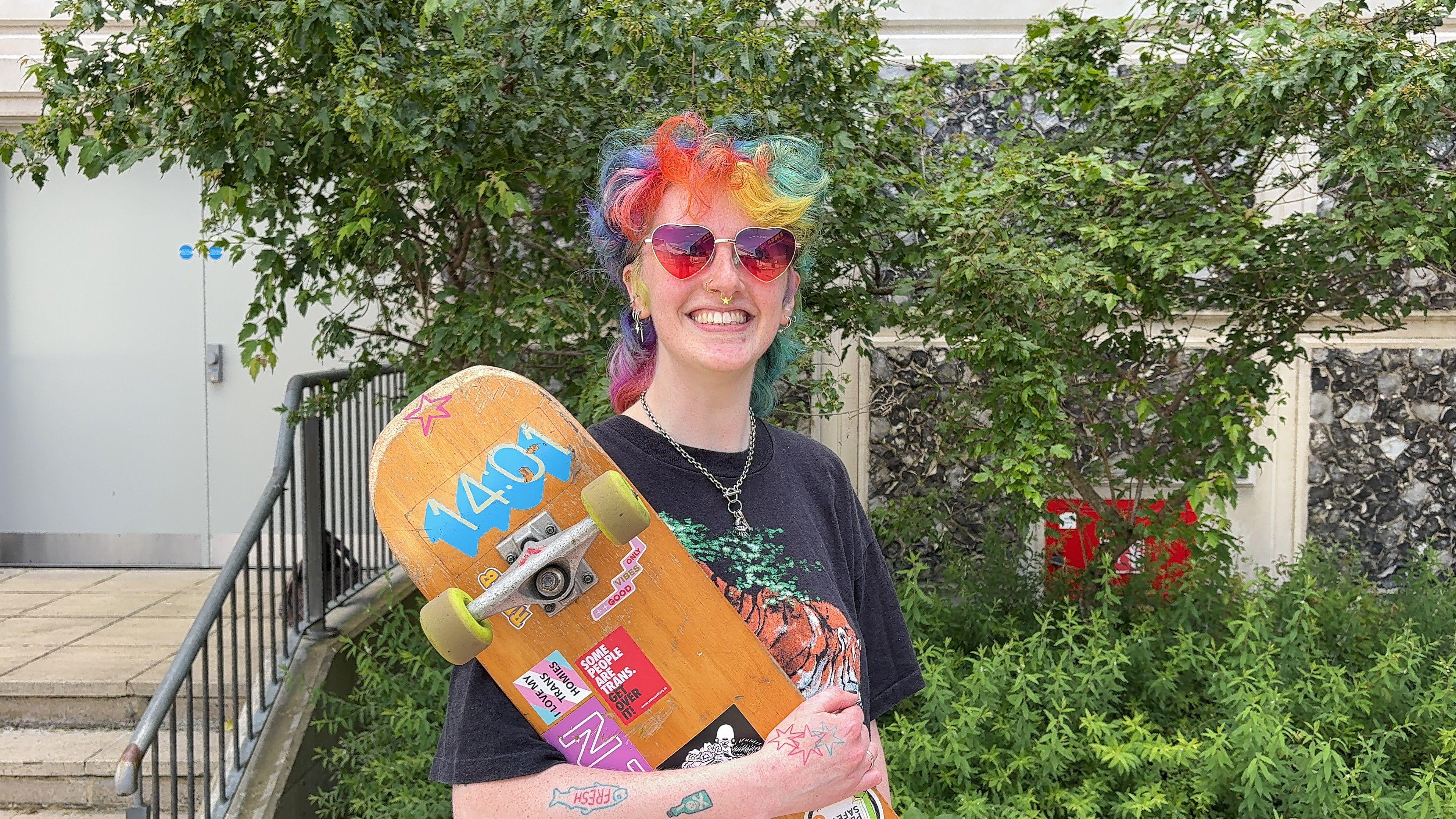Ozzy Crawley stood with their skateboard in their arms. They have multi-coloured hair and a chain around their neck. They are wearing heart-shaped sunglasses and have piercings on their face and tattoos on their arms.