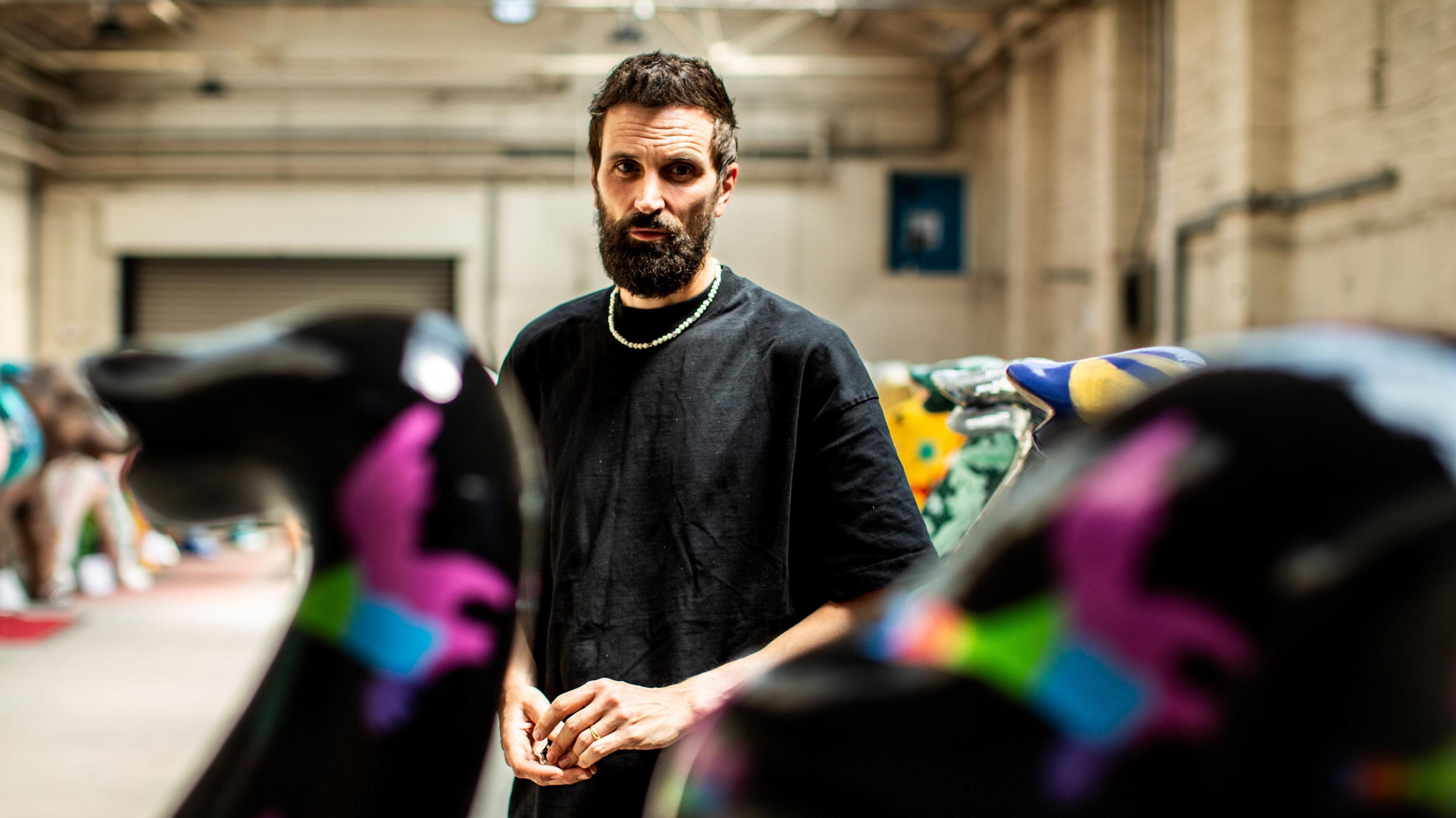 Serge Pizzorno wearing a black t shirt and chain, stood among colourfully painted elephant sculptures