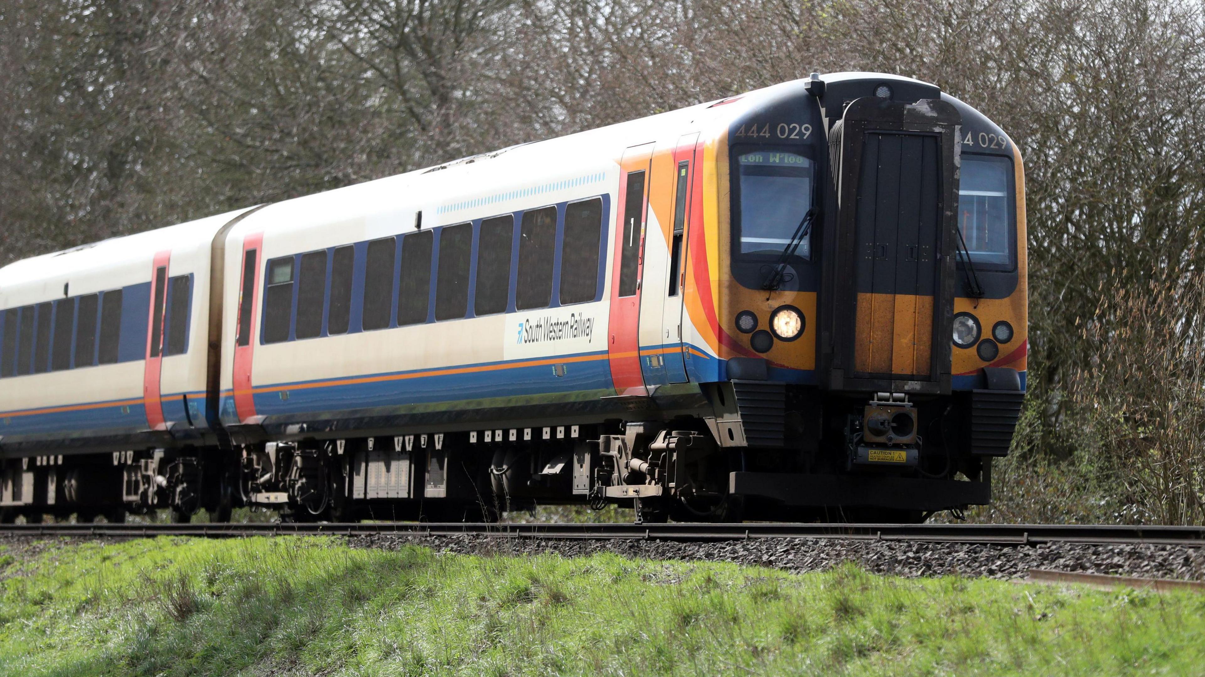 A South Western Railway train makes its way along the railway line near to Basingstoke in Hampshire. Green grass is seen either side and tall trees in the background.