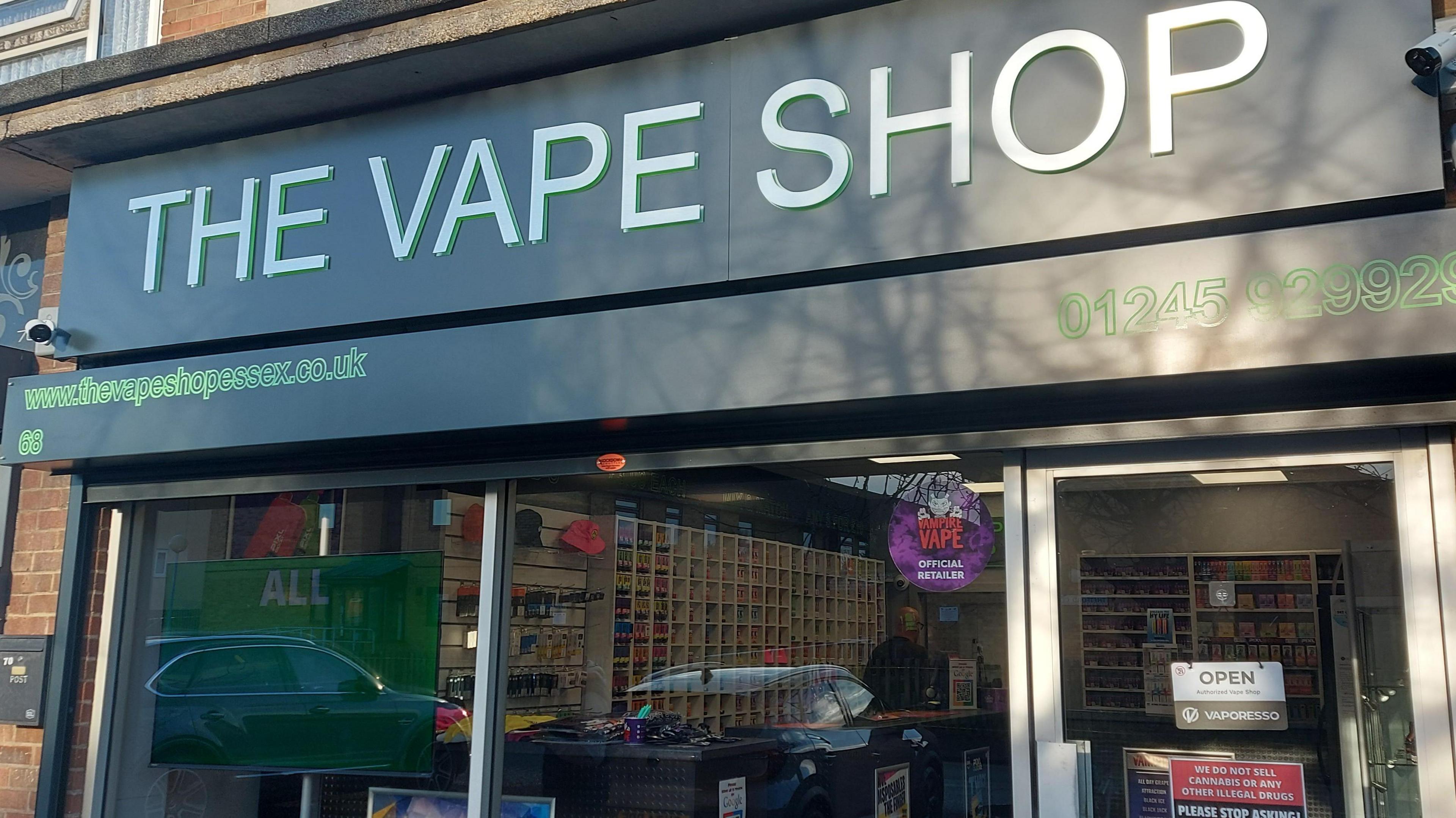 A shopfront saying "the vape shop" in bold white lettering, with shelves full of vapes visible through the glass and a sign saying the shop is open.