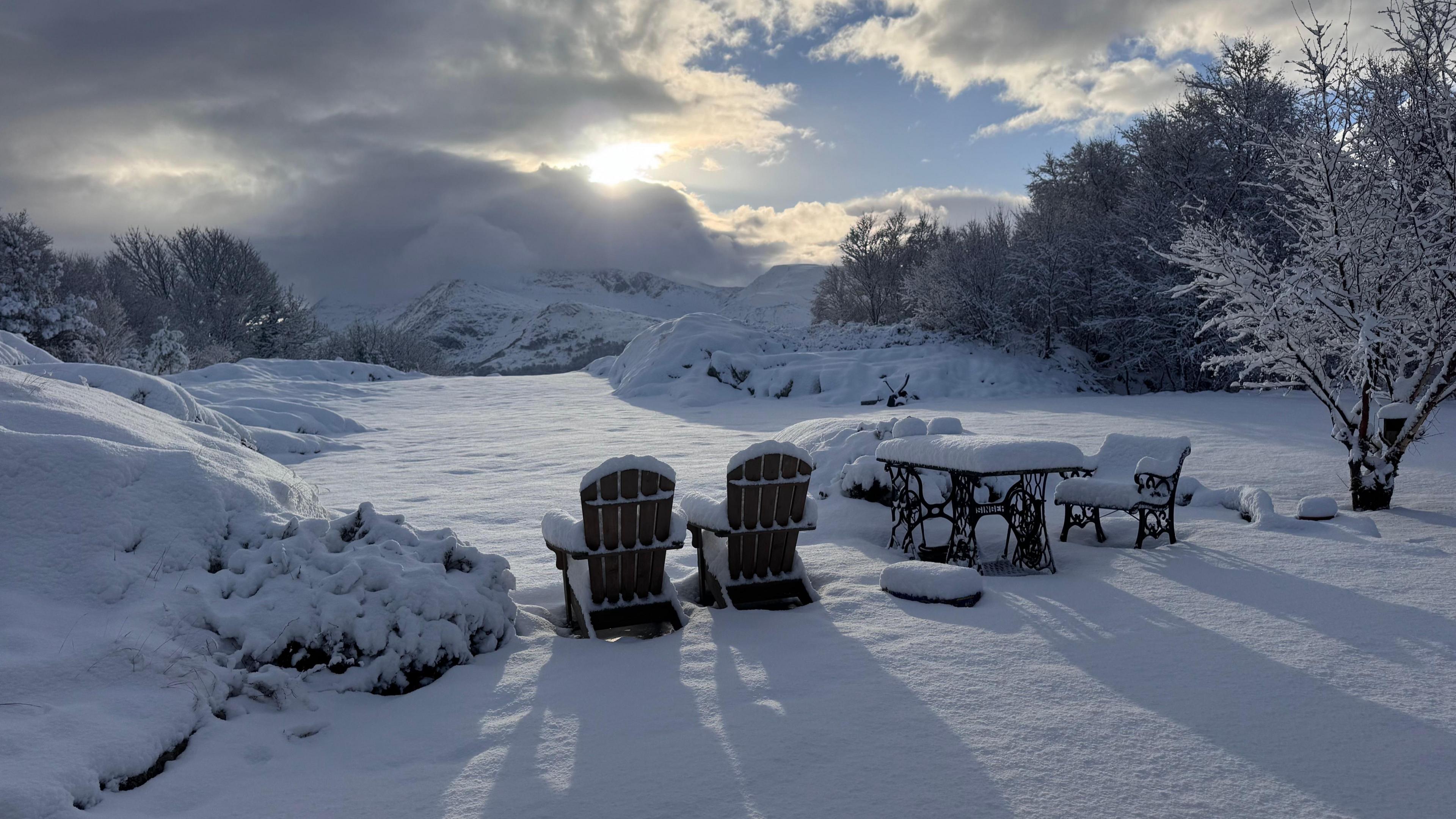 Large garden surrounded by shrubbery with a table and three chairs covered in deep snow, snow-covered hills in the distance with sun poking through thick cloud 