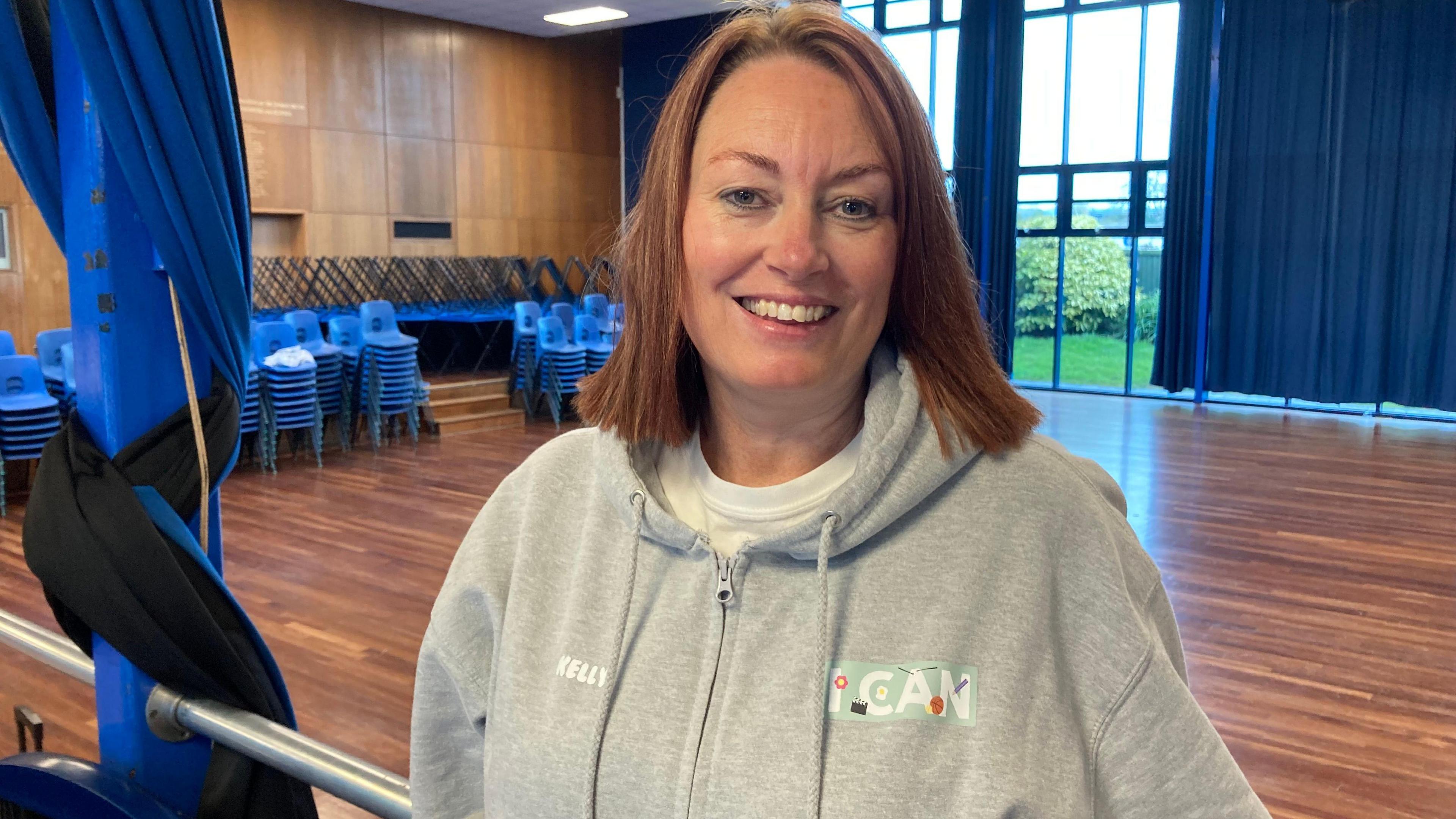 Kelly Cornish is standing in a rehearsal space with stacked chairs in the background. She is wearing a grey hoodie and is smiling at the camera.