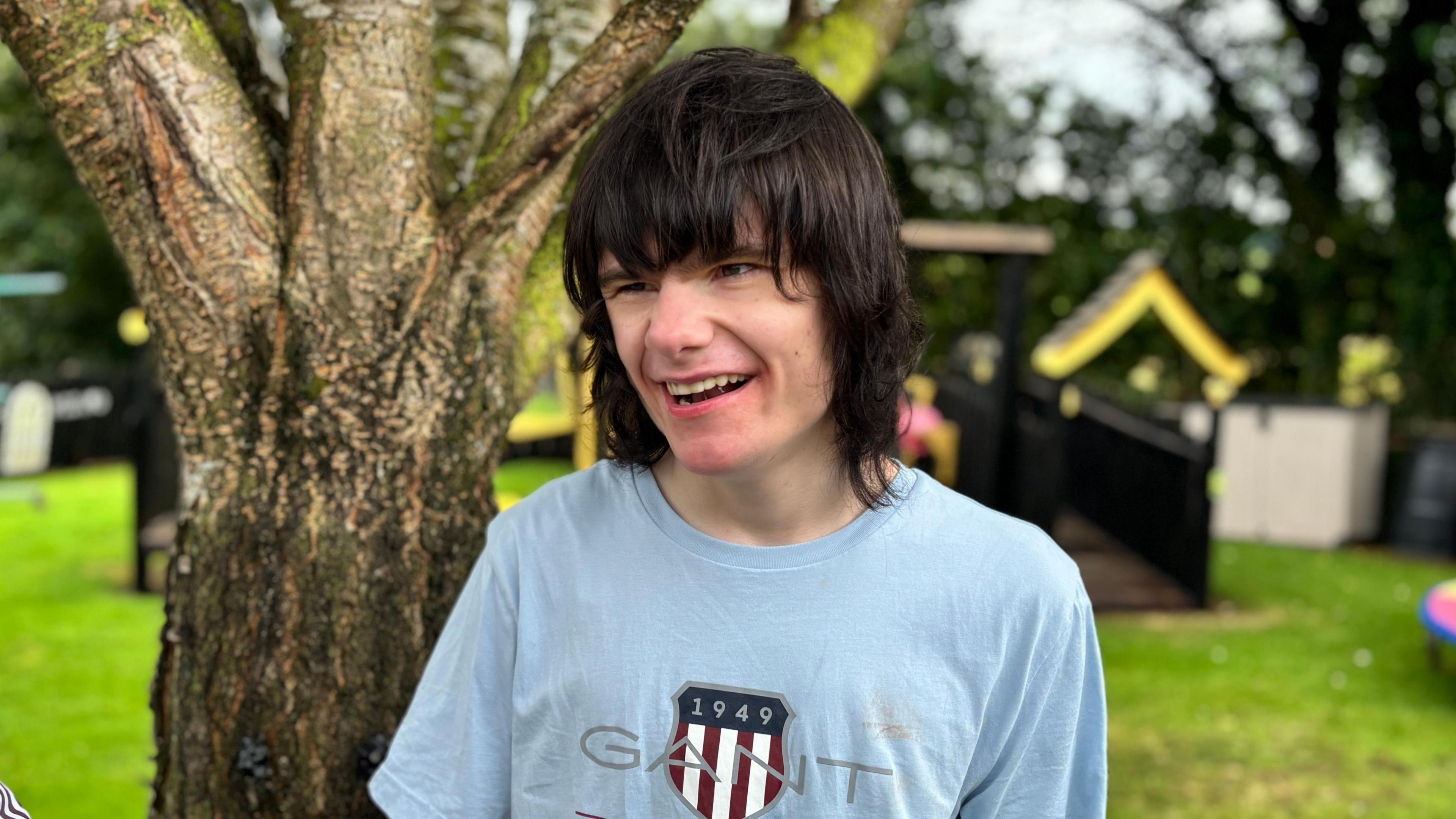 A young man stands in a garden. He has dark shoulder length hair with a heavy fringe. He is smiling widely and wearing a blue t-shirt. behind him is a bright sunny garden, with grass and treats and some play equipment. 