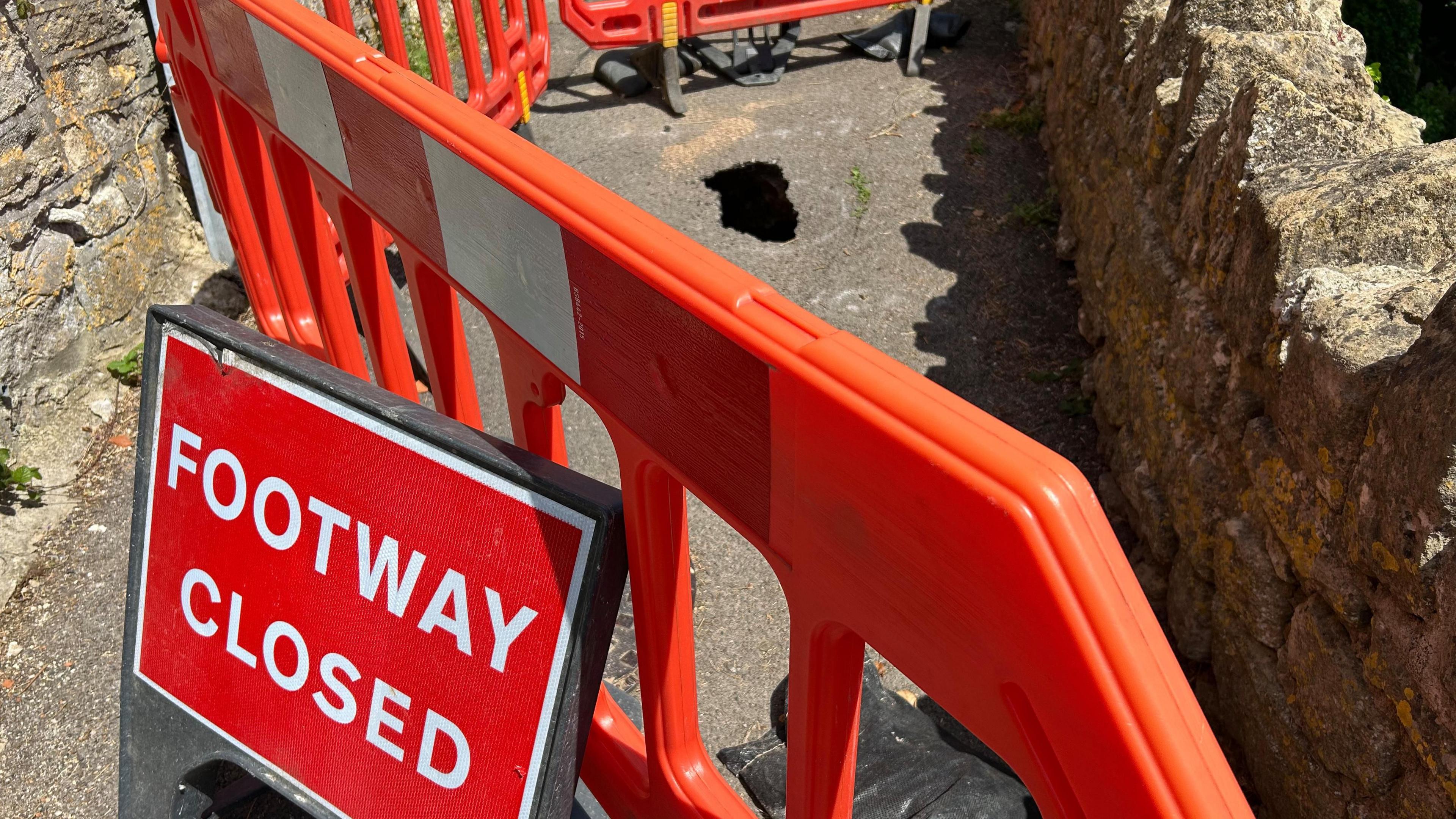 A red footway closed sign next to some bright orange temporary fencing. Behind the fencing a hole can be seen in a footpath.