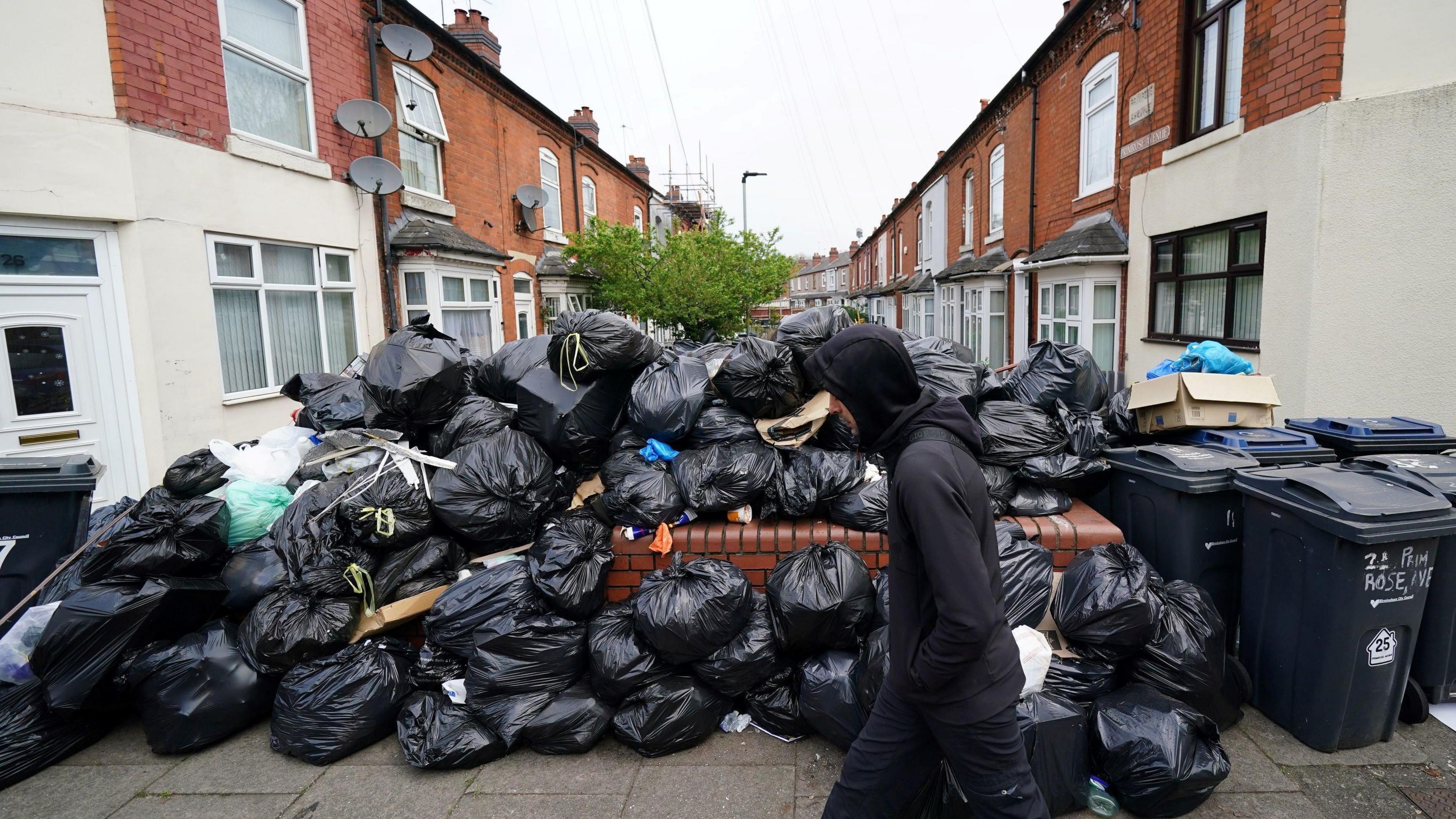 A man in a black hoodie walks past black bin bags piled up on a residential street, with terraced houses on either side.