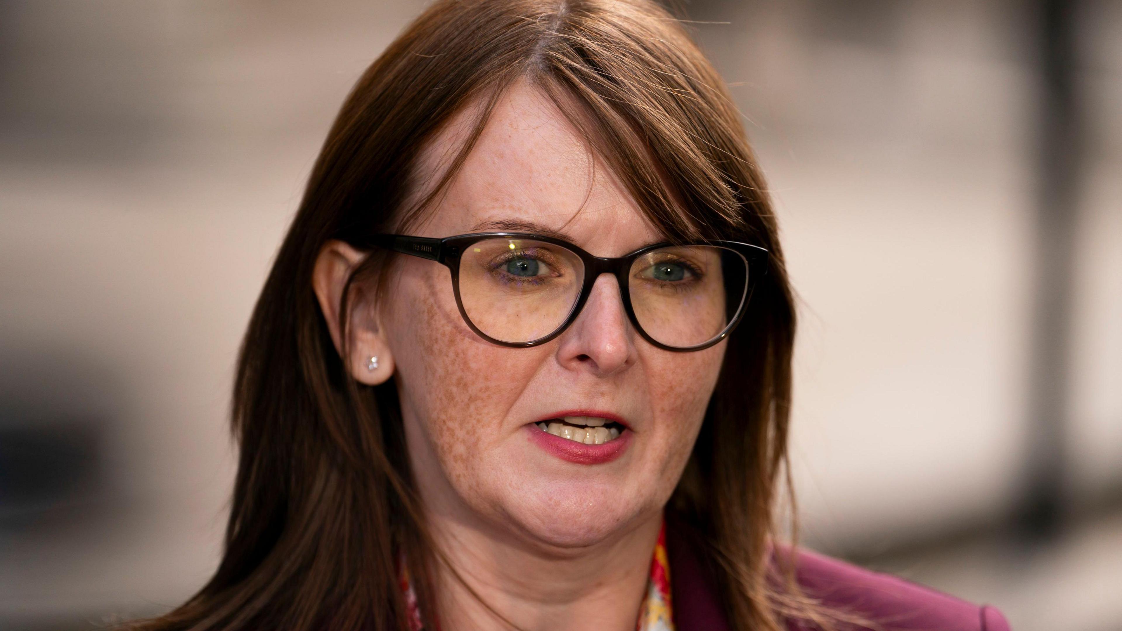 Caoimhe Archilbald is wearing black glasses and a burgundy jacket, she has shoulder-length auburn hair, pink lipstick and has a diamond stud earring in her right ear