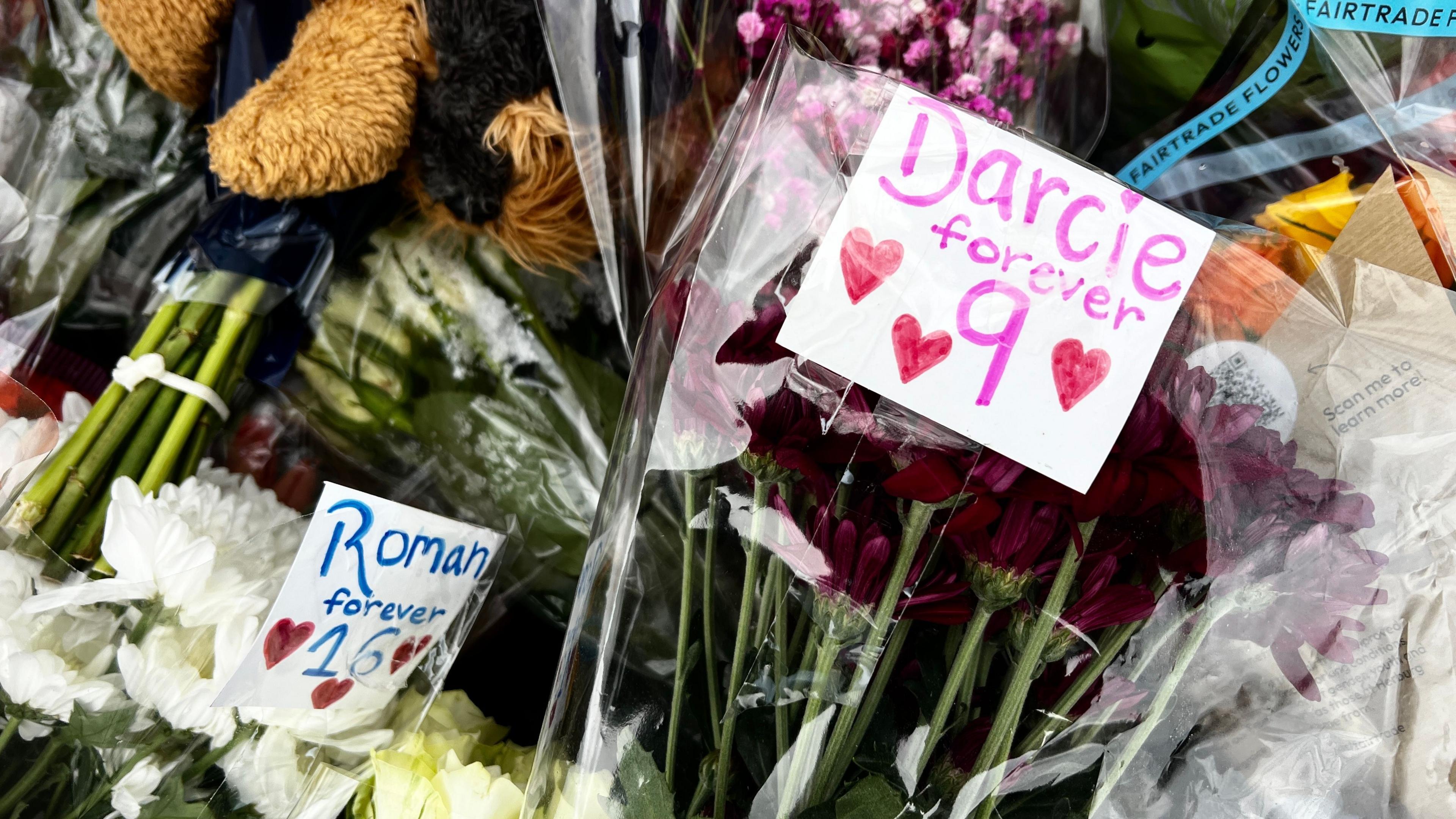 Bunches of flowers piled on top of each other. Two at the forefront of the picture have signs that read "Roman forever 16" and "Darcie forever nine" respectively.
