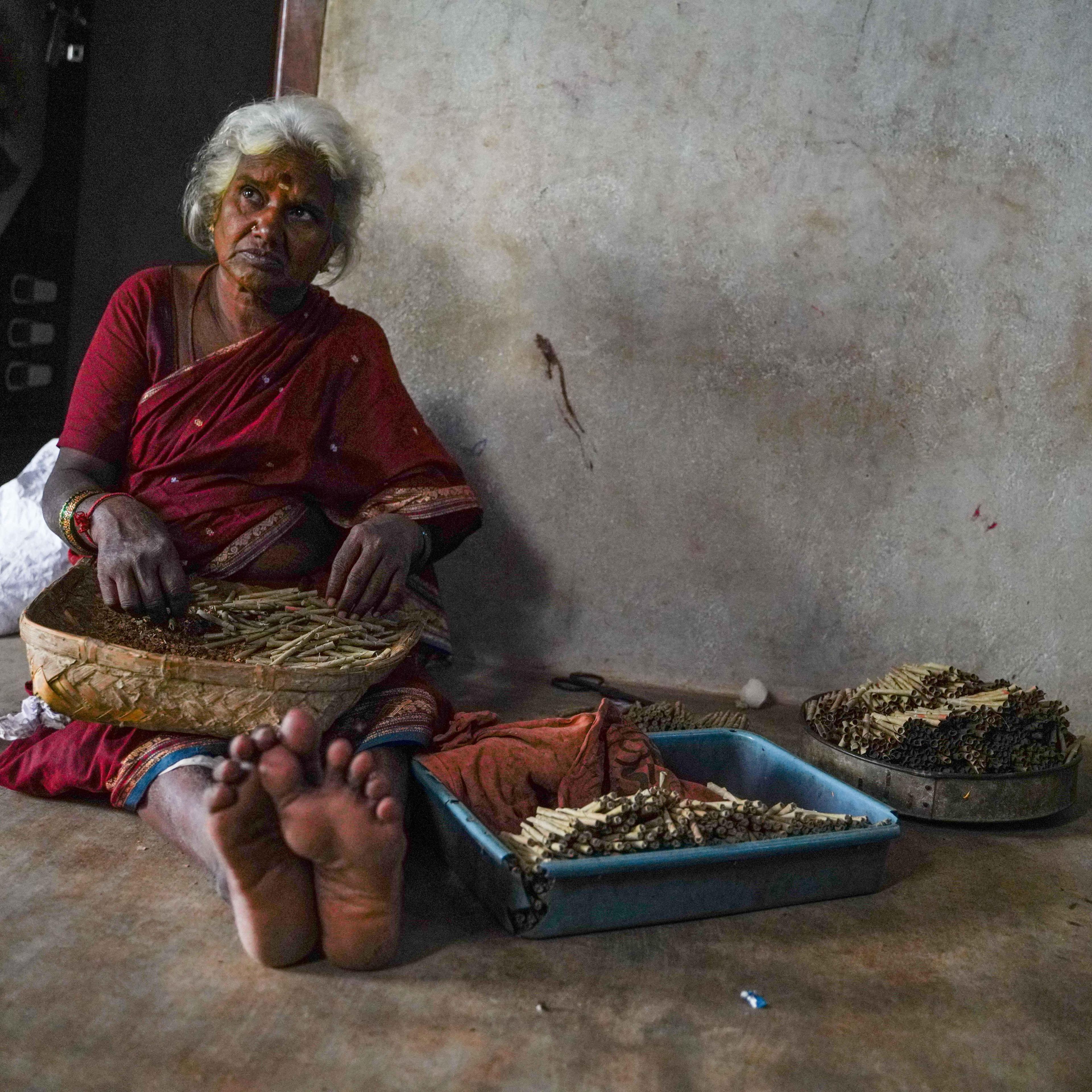 An elderly woman wearing a red saree with golden borders sits with a basket of hand-rolled cigarettes. There are two other stacks of rolled beedis placed next to her. 