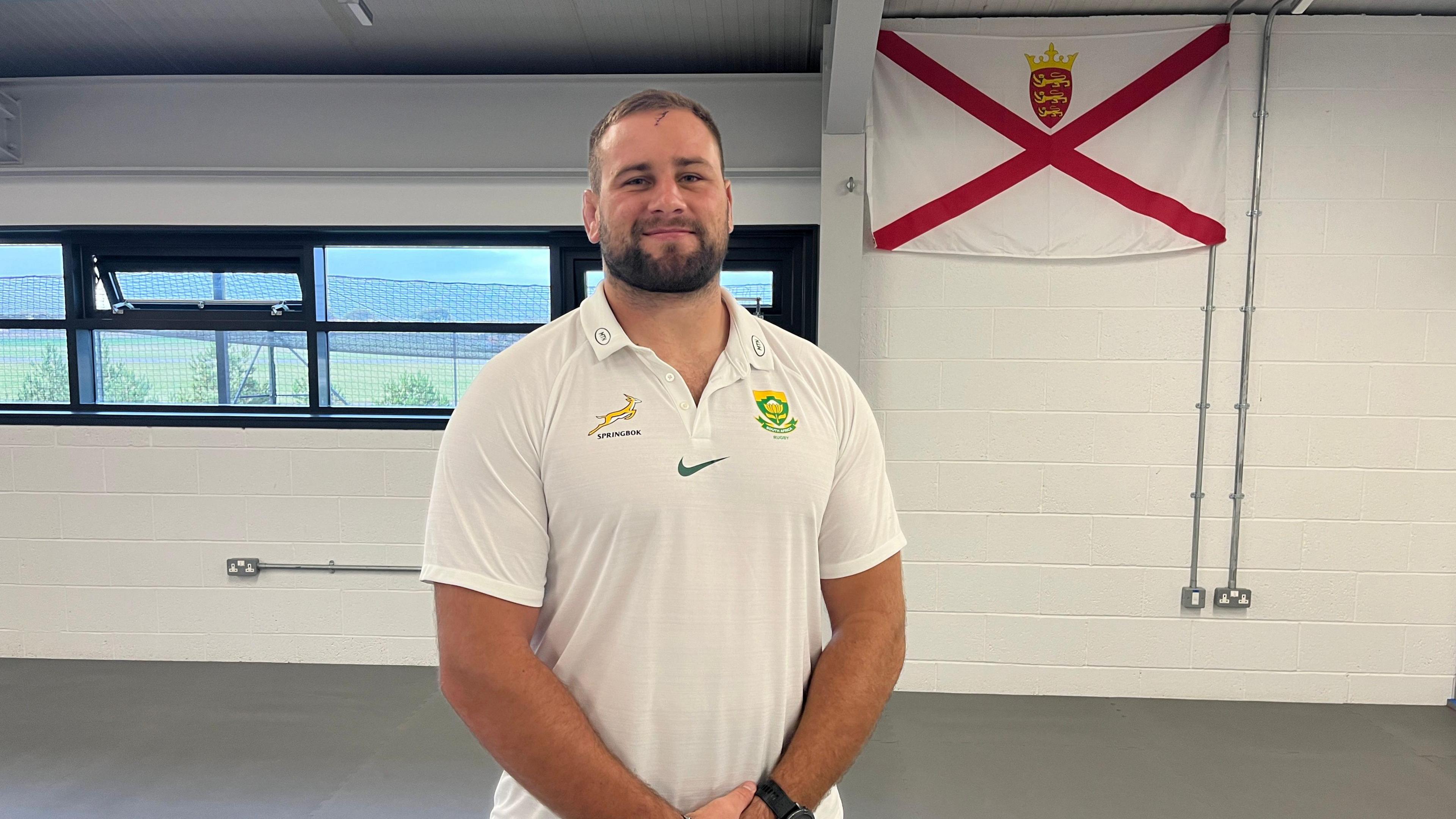 Thomas du Toit wearing a white T-shirt with the Springbok logo on the left, a Nike tick at the centre and a badge on the right. He is stood with his hands together in front of him and is smiling at the camera. In the background on the right is a Jersey flag, with the logo at the top and windows looking over the green pitch on the left. 