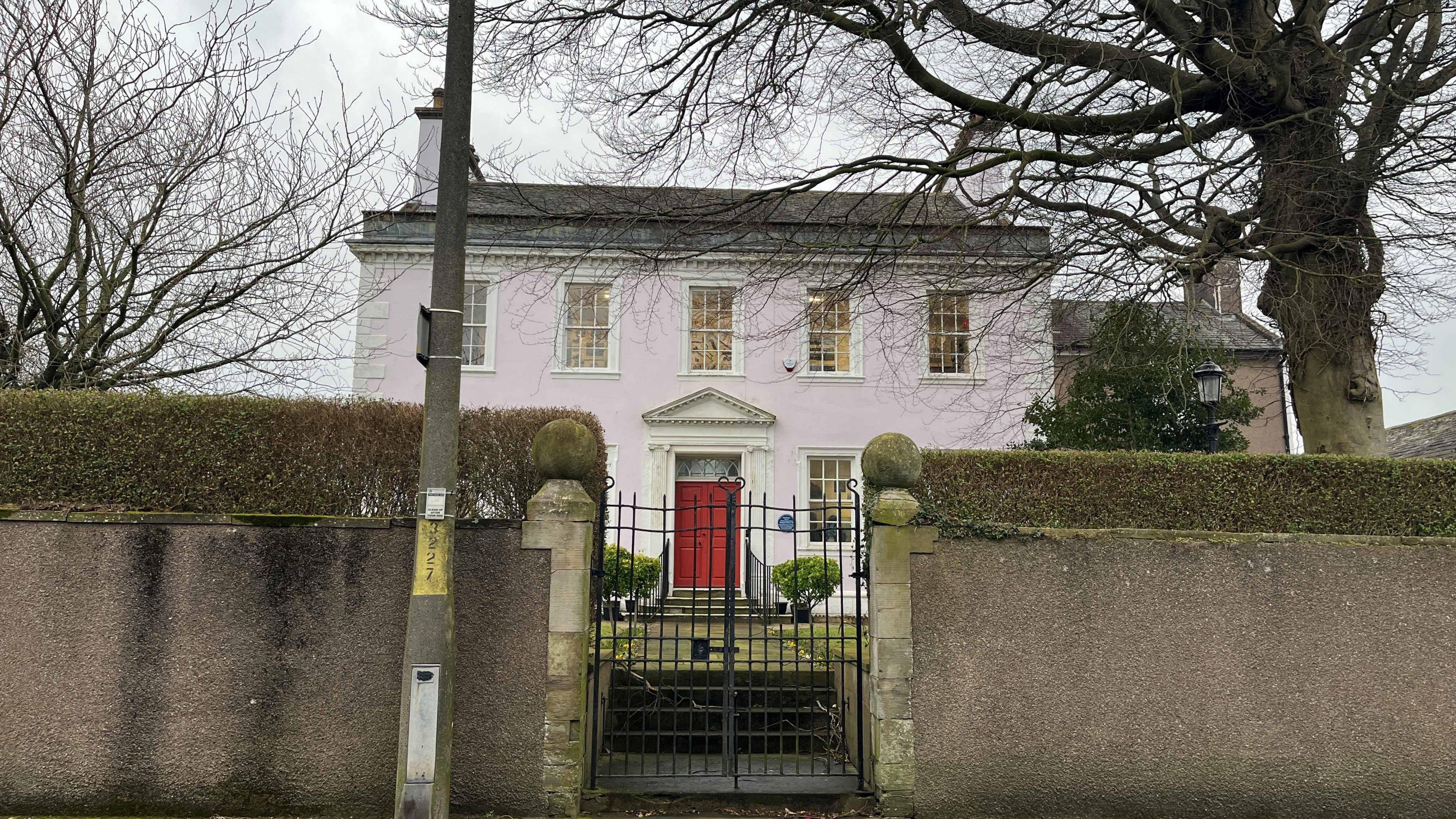 A general view of the Helena Thompson museum. The Georgian house is painted pink with paned windows and a red front door. There is a small front garden with trees and a path leading from the wrought iron gate to the front door.