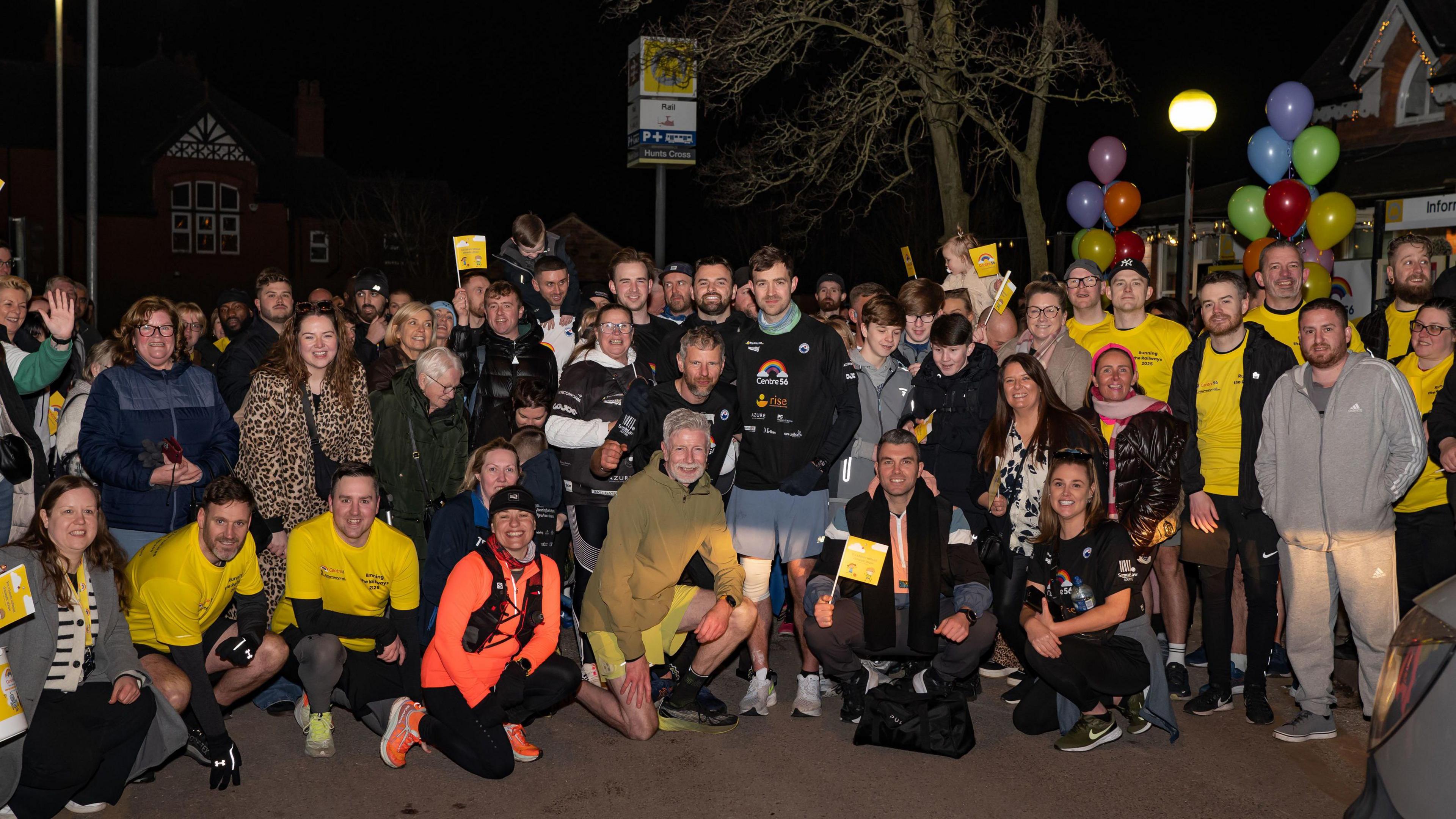 Alex Rigby, pictured in a black hoodie and surrounded with supporters after reaching Hunts Cross station - the final stop of his running challenge. The crowd have flags and balloons.