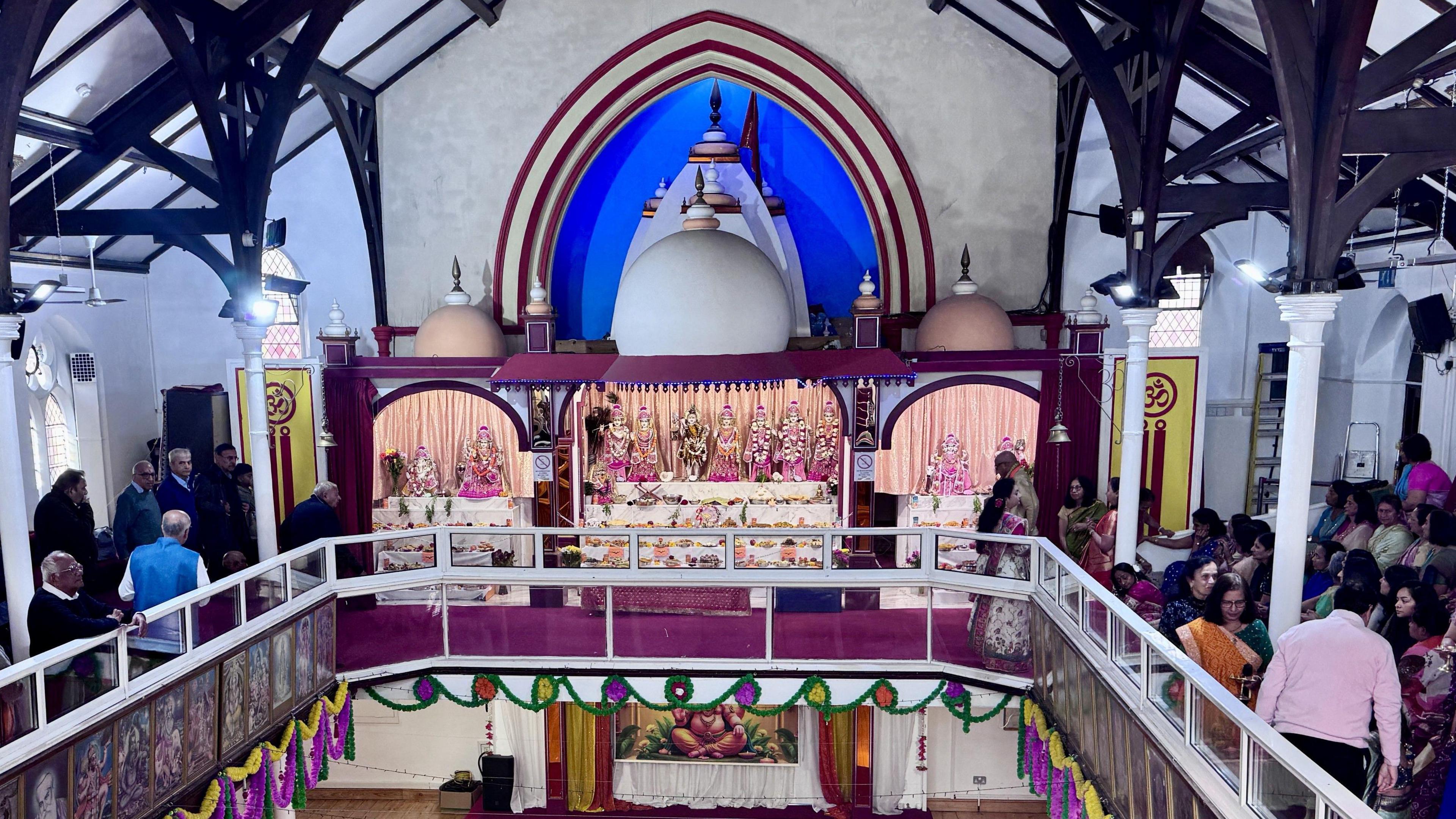 The image is a view of the inside of a Hindu temple, you can see the balcony area which has people sat around it. At the end of the balcony area there is a colourful display of the deities in the Main Sanctum of the Hindu Temple. He is wearing a gold traditional Hindu garment with orange material draped around his neck.