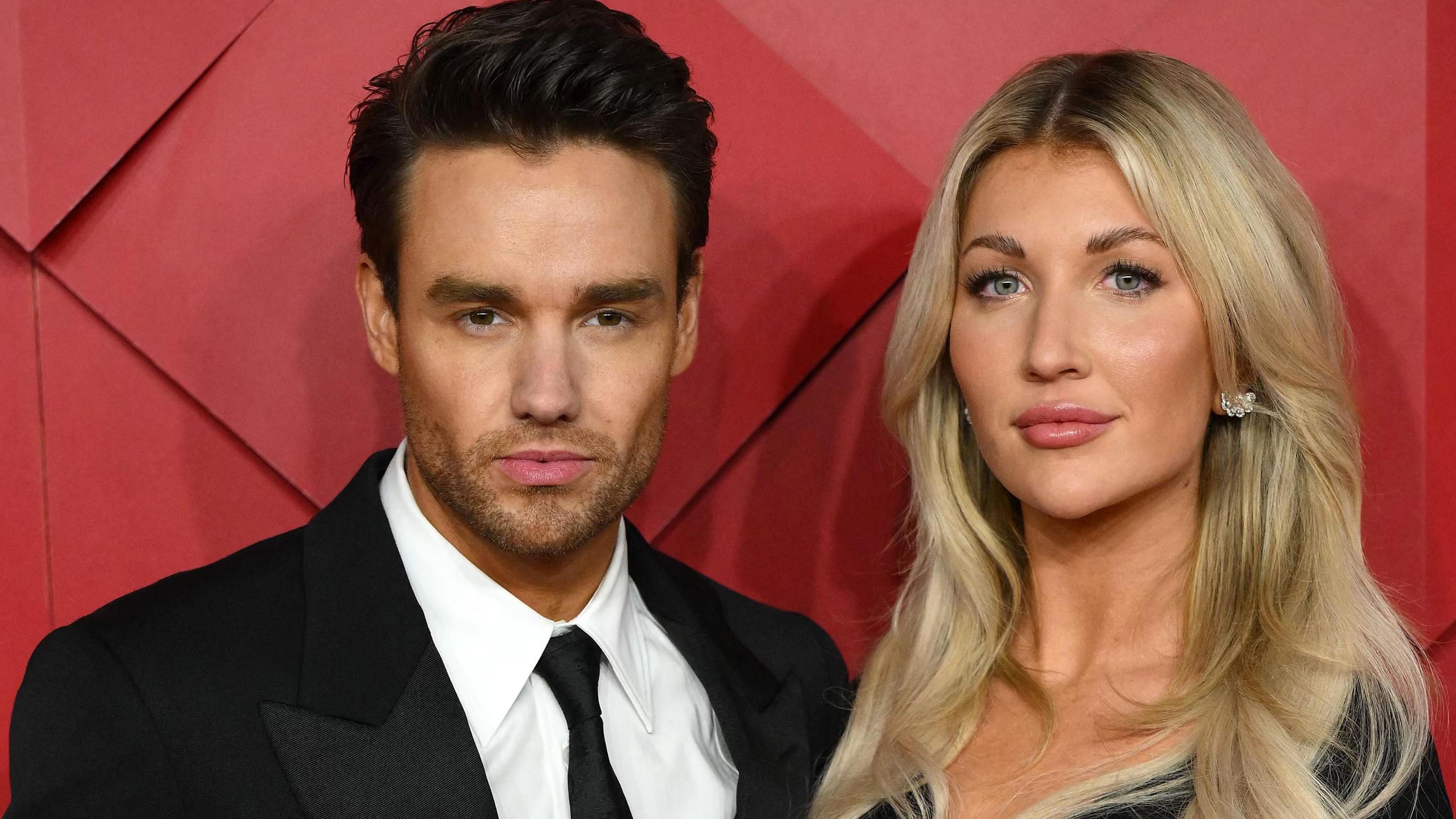 Liam Payne in suit and tie stands beside a smiling Kate Cassidy, with blonde hair and glittery earrings, against a red background