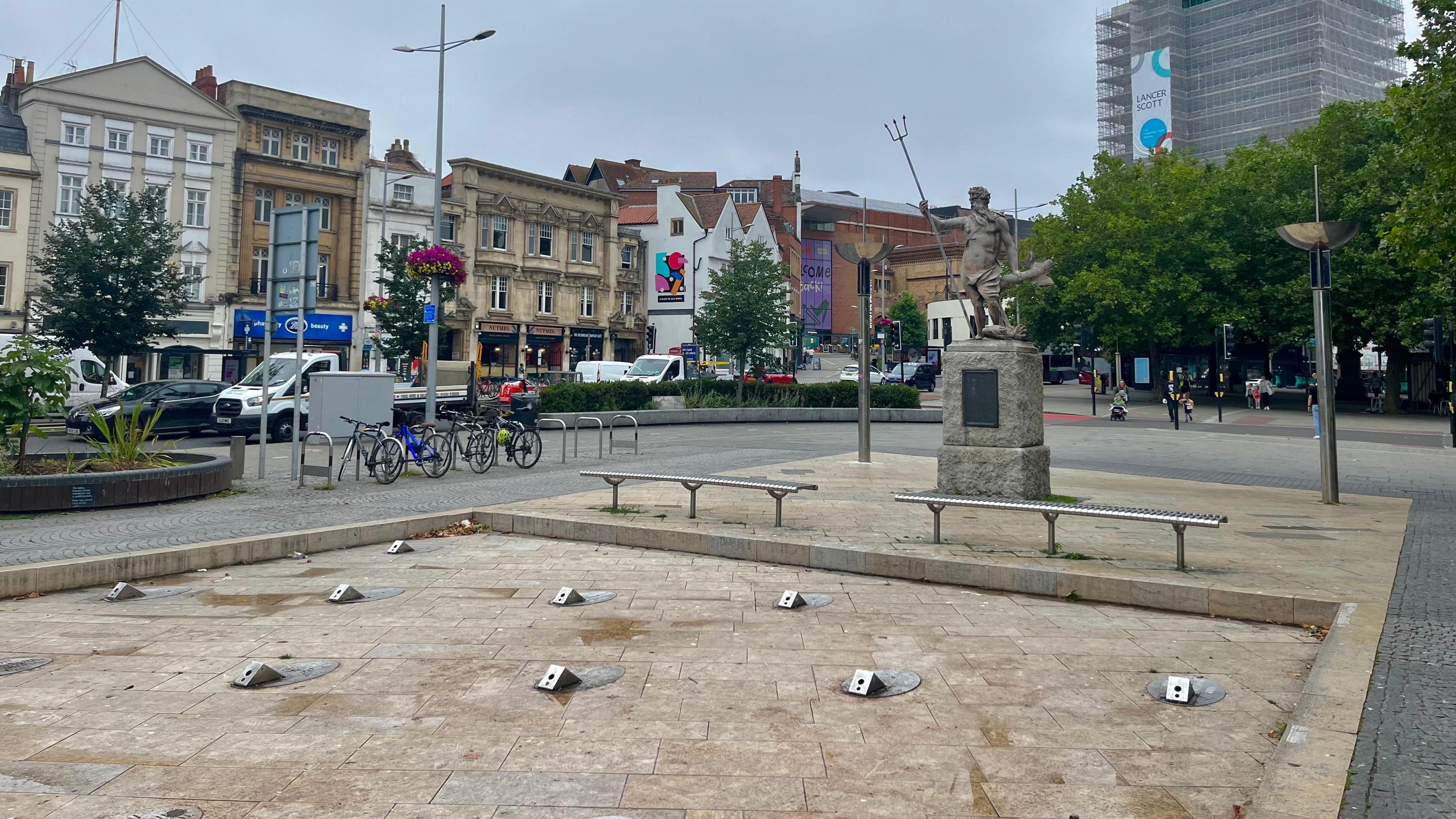 Bristol City Centre fountains. It is a large, square, slightly sunken structure with small silver fountain nibs embedded in the stone and benches around the outside. There is no water flowing in the fountain.