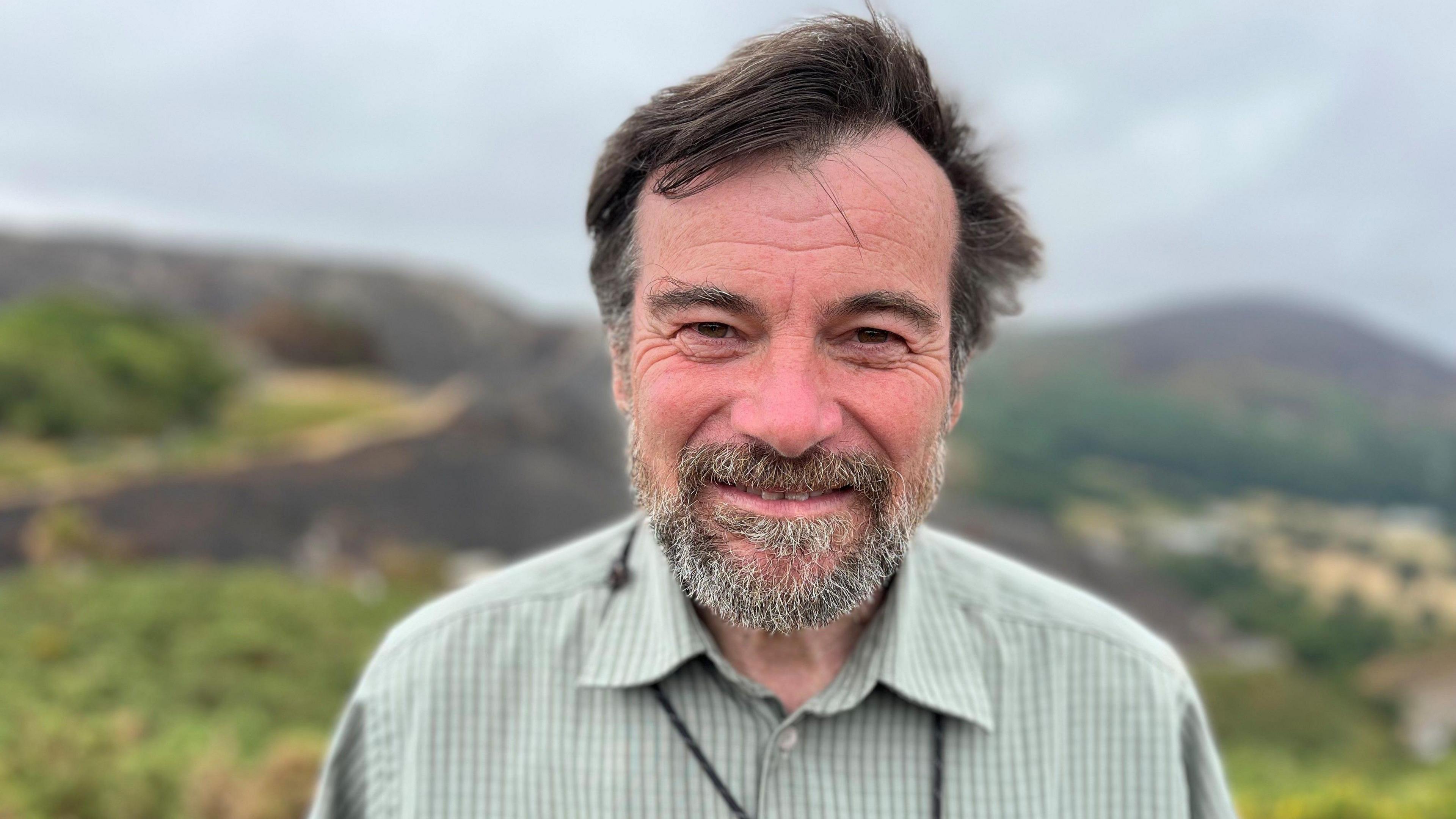 A picture of Julian Thompson looking at the camera. He is stood in the Pensychnant Conservation Centre and is wearing a green and white chequered shirt.