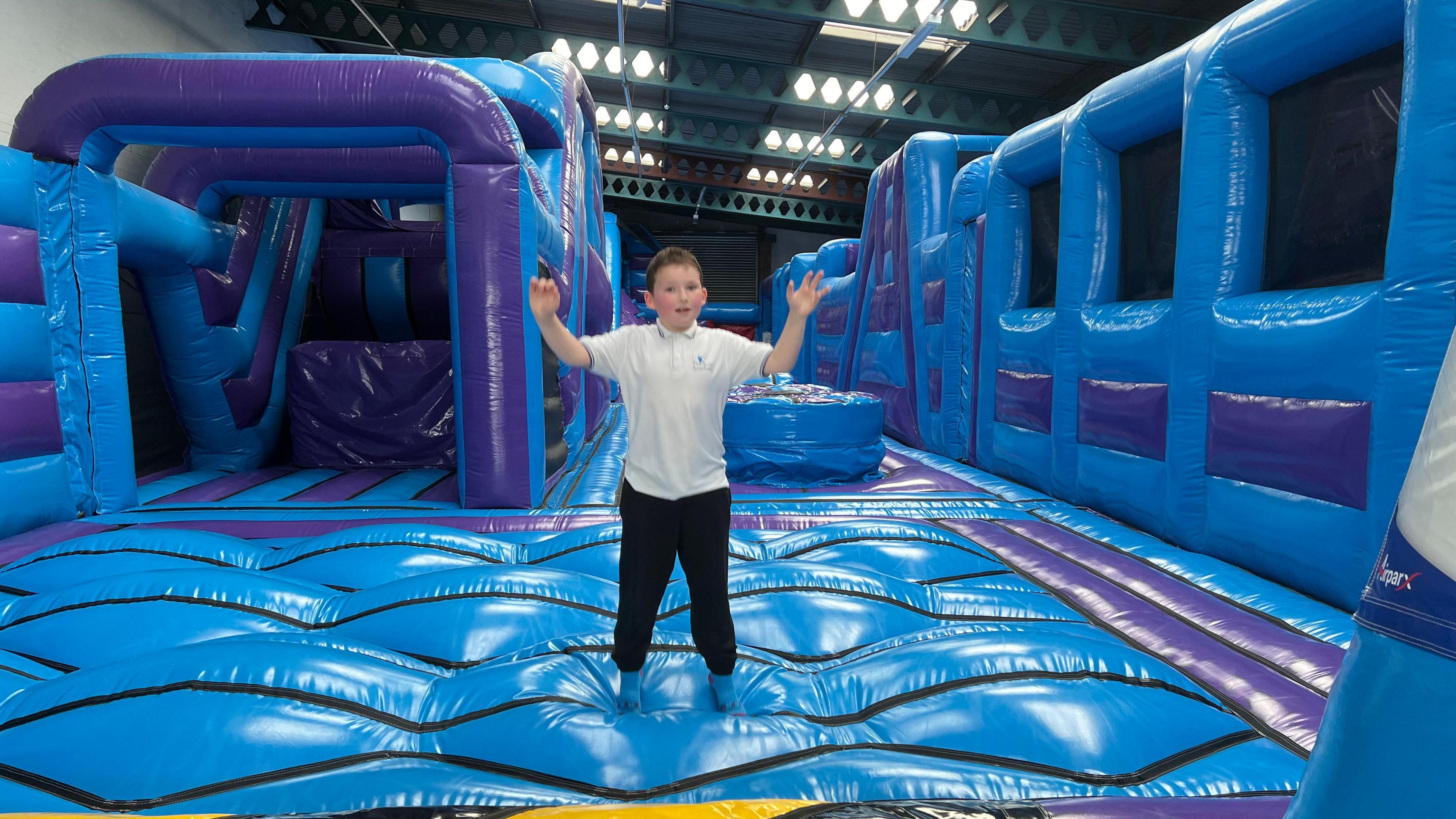 Ted wearing black tracksuit bottoms and a white polo shirt which says 'Venturers' Academy' on it. He is standing in the middle of a blue inflatable obstacle course with both arms in the air.