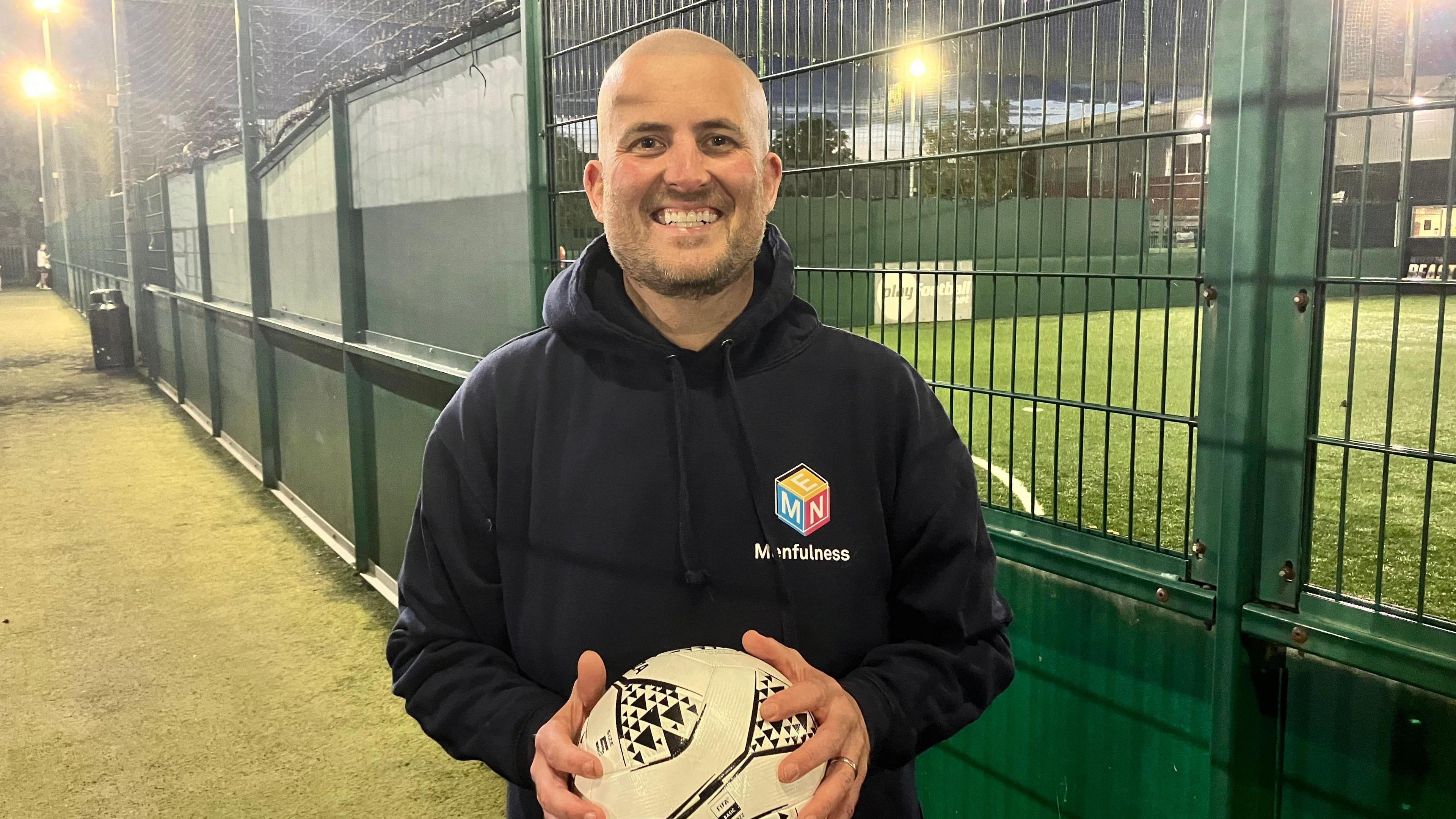 Jack Woodhams, chief executive of York mental health charity Menfulness, is smiling at the camera. He is wearing a black hoodie, holding a football and is standing in front of five-a-side pitches.