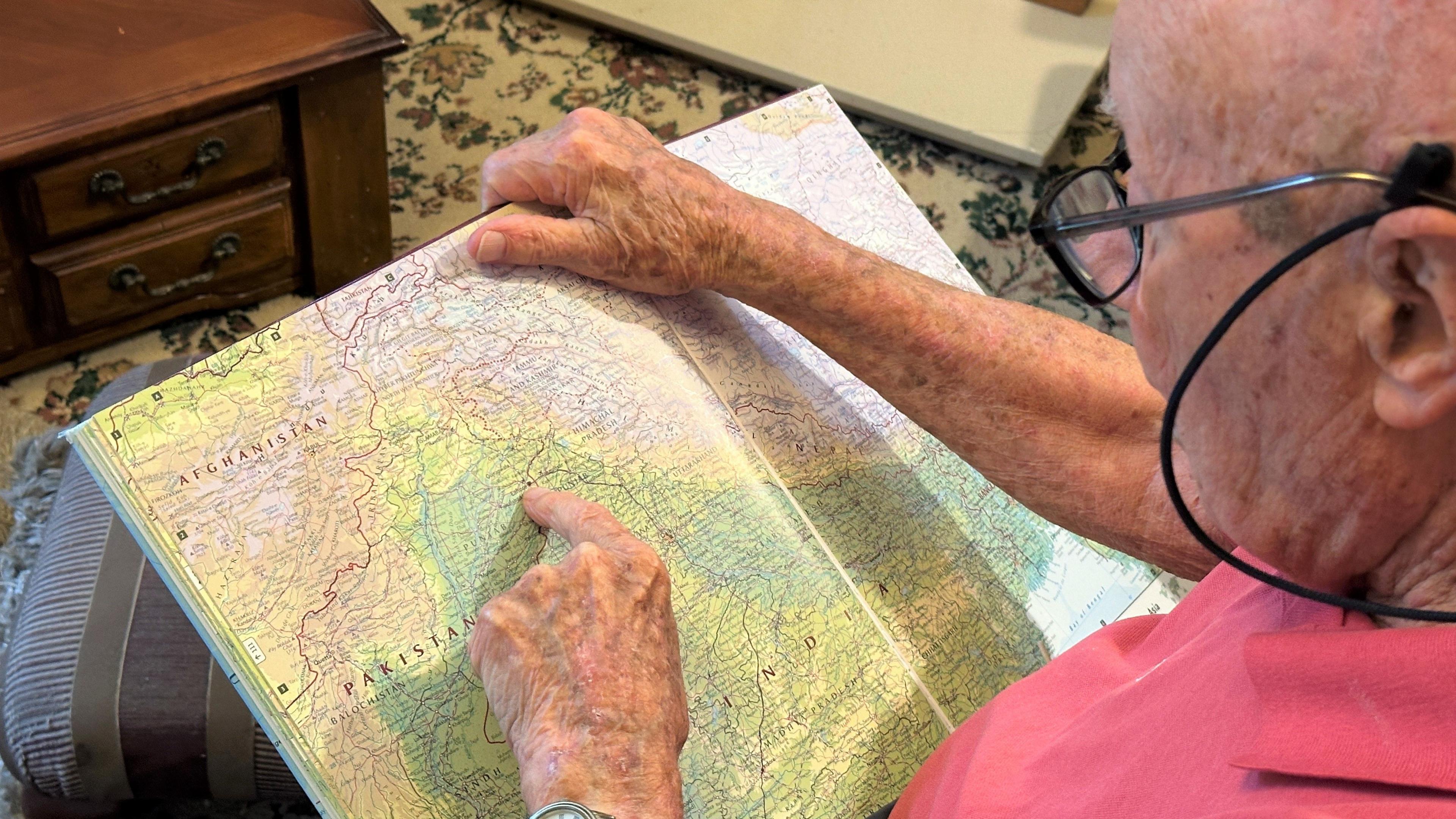 Peter points to Lahore on an atlas, remembering the warm hospitality him and his friends received. Peter is wearing a red t-shirt and glasses in his living room, which has a patterned carpet.