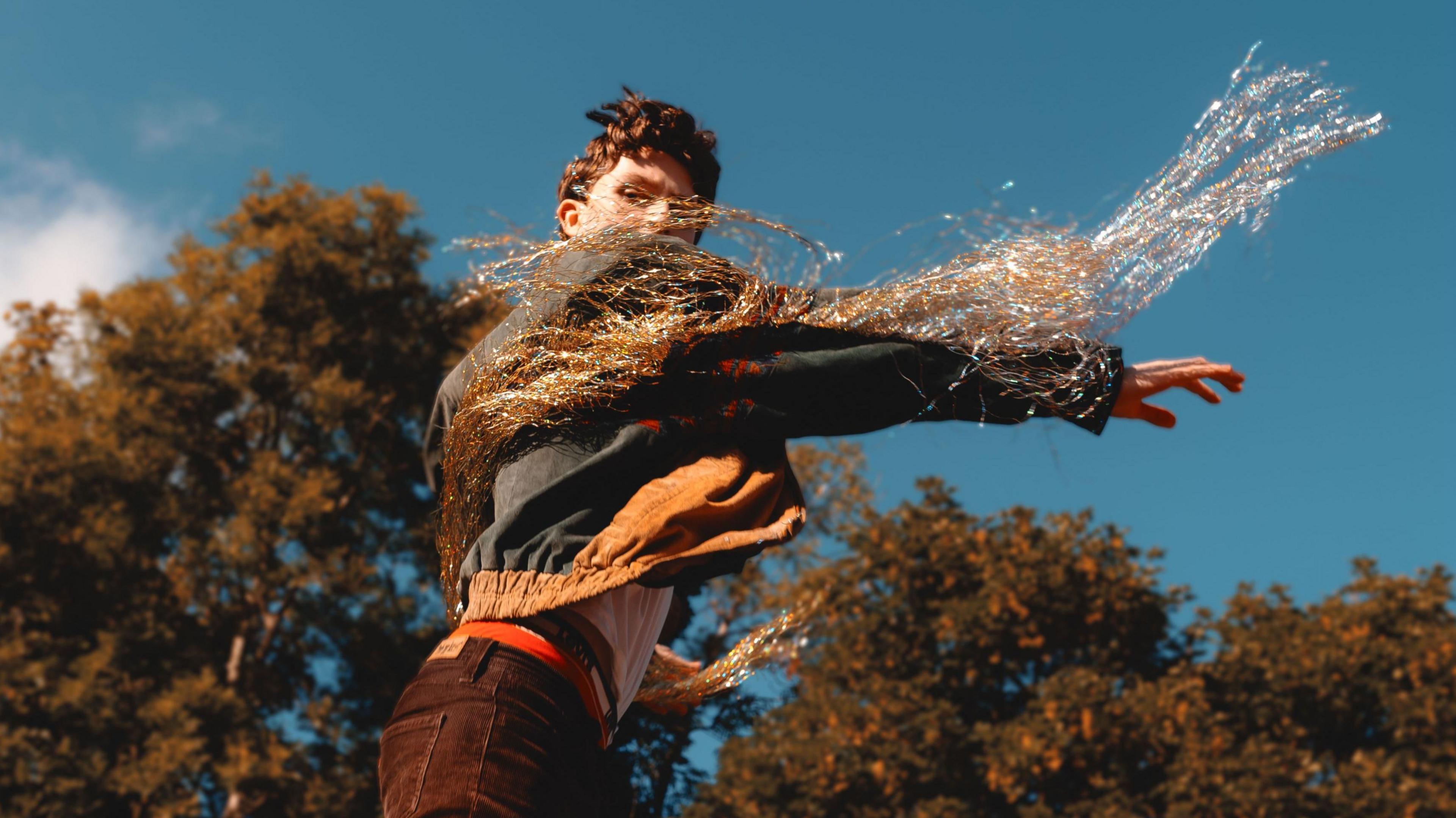 A man wearing a blue and burnt orange jacket with lots of dramatic fringing, white t-shirt, and brown corduroy trousers stands in front of a row of trees. The sky is blue with very few clouds.