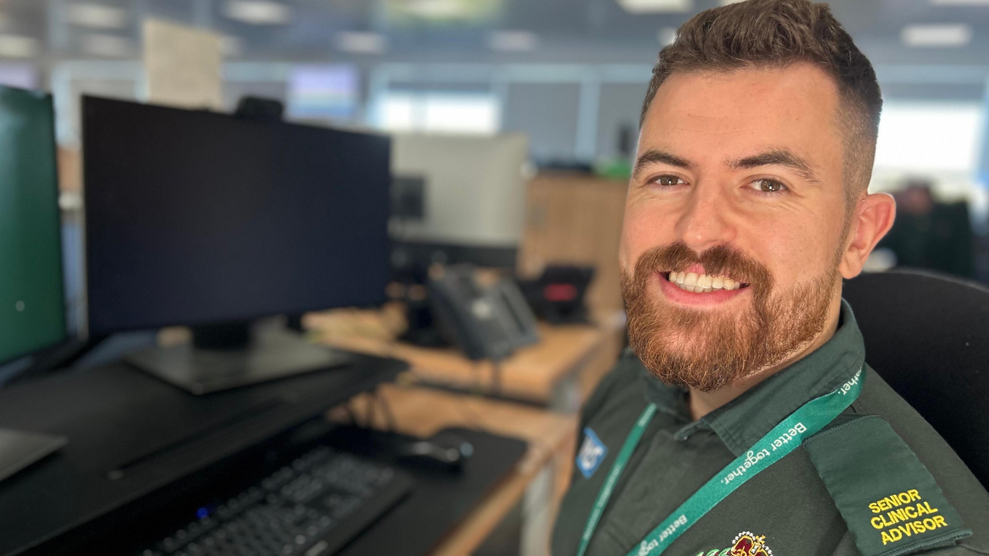 Kyle is pictured wearing his dark green North East Ambulance Service uniform. He has dark brown hair and a goatee beard. 