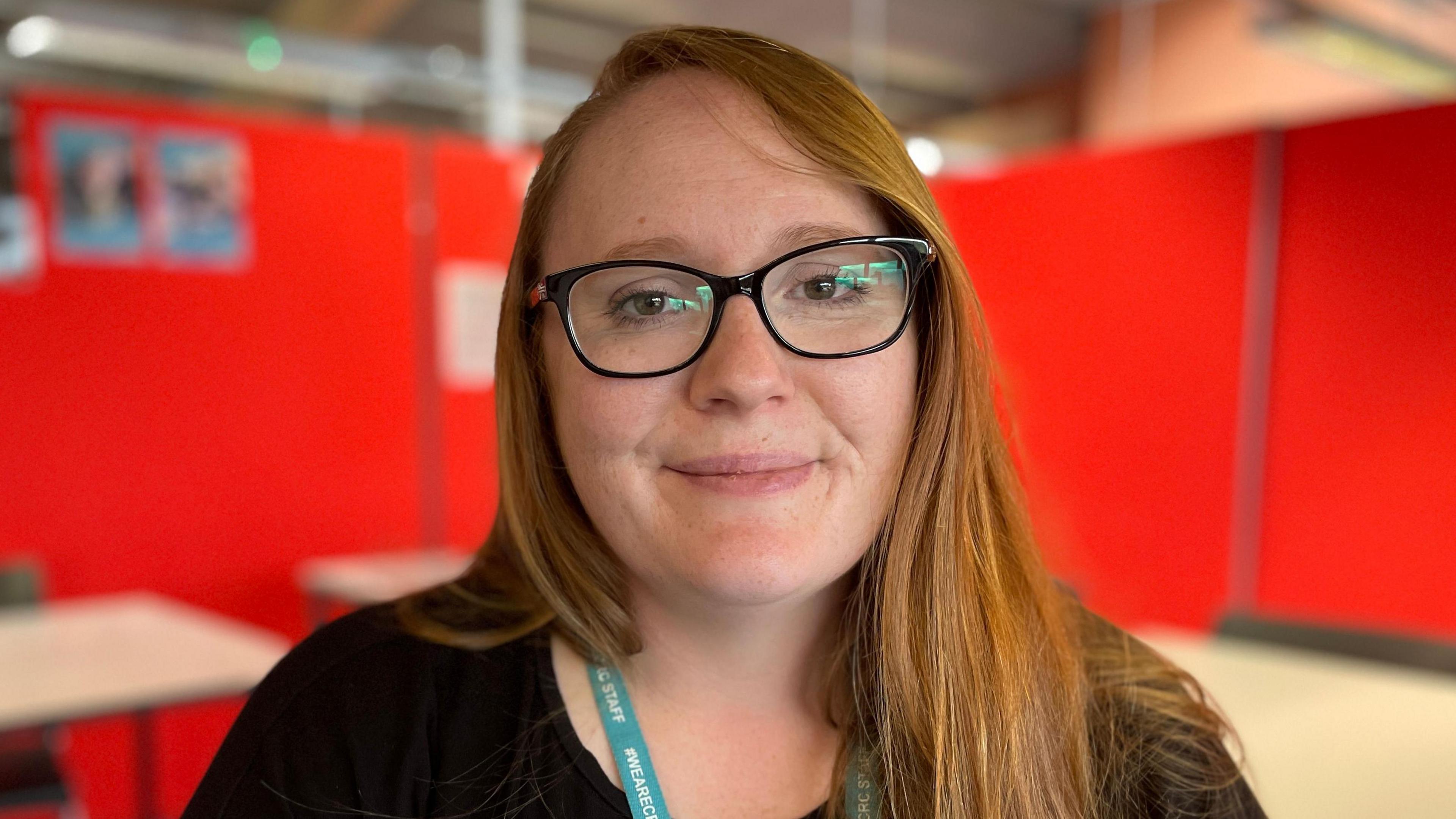 Laurie is wearing black framed glasses, a black top and green lanyard. She has red hair and she's standing in a computer room surrounded by red boards.