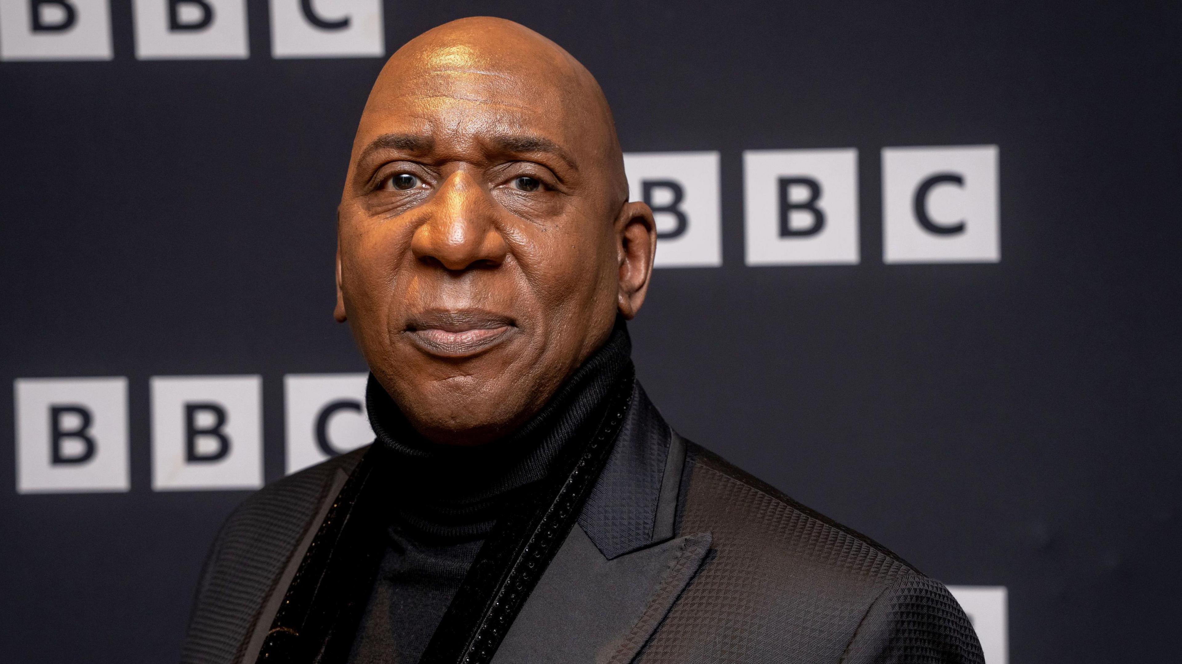 The actor wears a black polo neck jumper under a black suit and stands in front of a screen with the BBC logo on it.