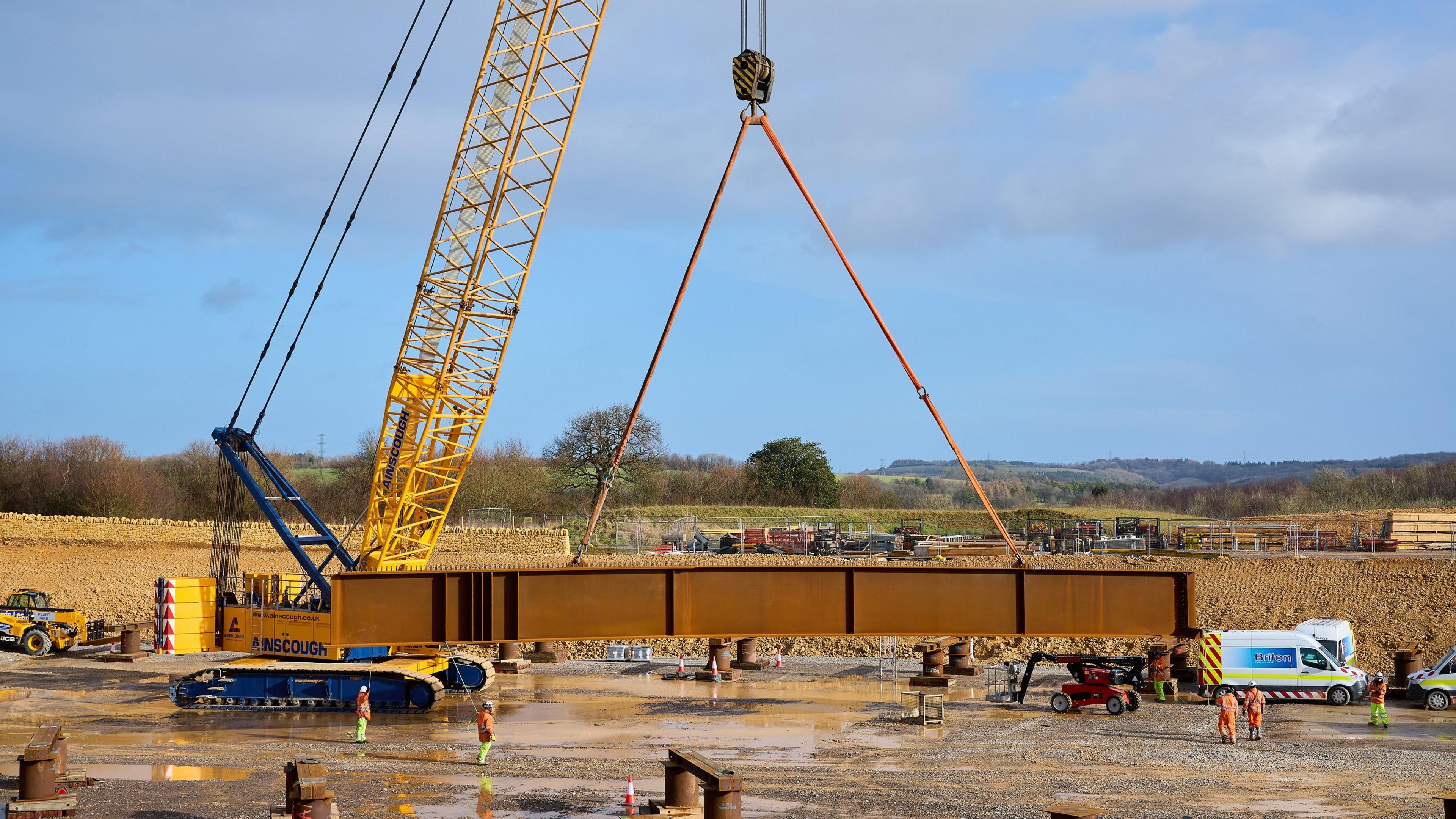 A yellow and blue crane holding up a large copper coloured beam which is suspended in mid air. Below it are five workers, dressed in orange and yellow hi-vis, who are navigating the lowering of the beam. It is a sunny day with blue skies and wispy clouds. 