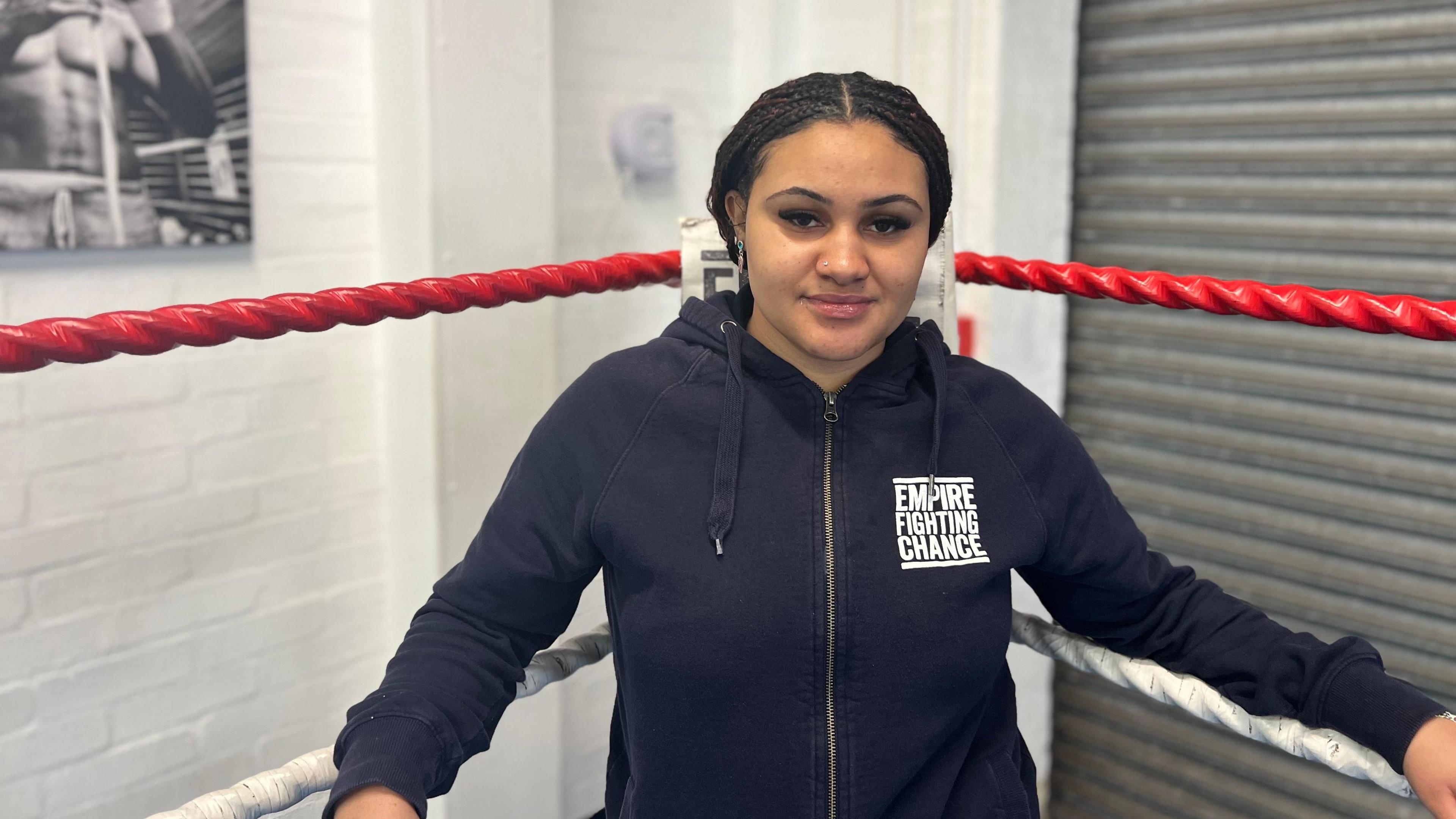 Serena is standing in a boxing ring. She is wearing a blue hoodie which says Empire Fighting Chance on the front. 