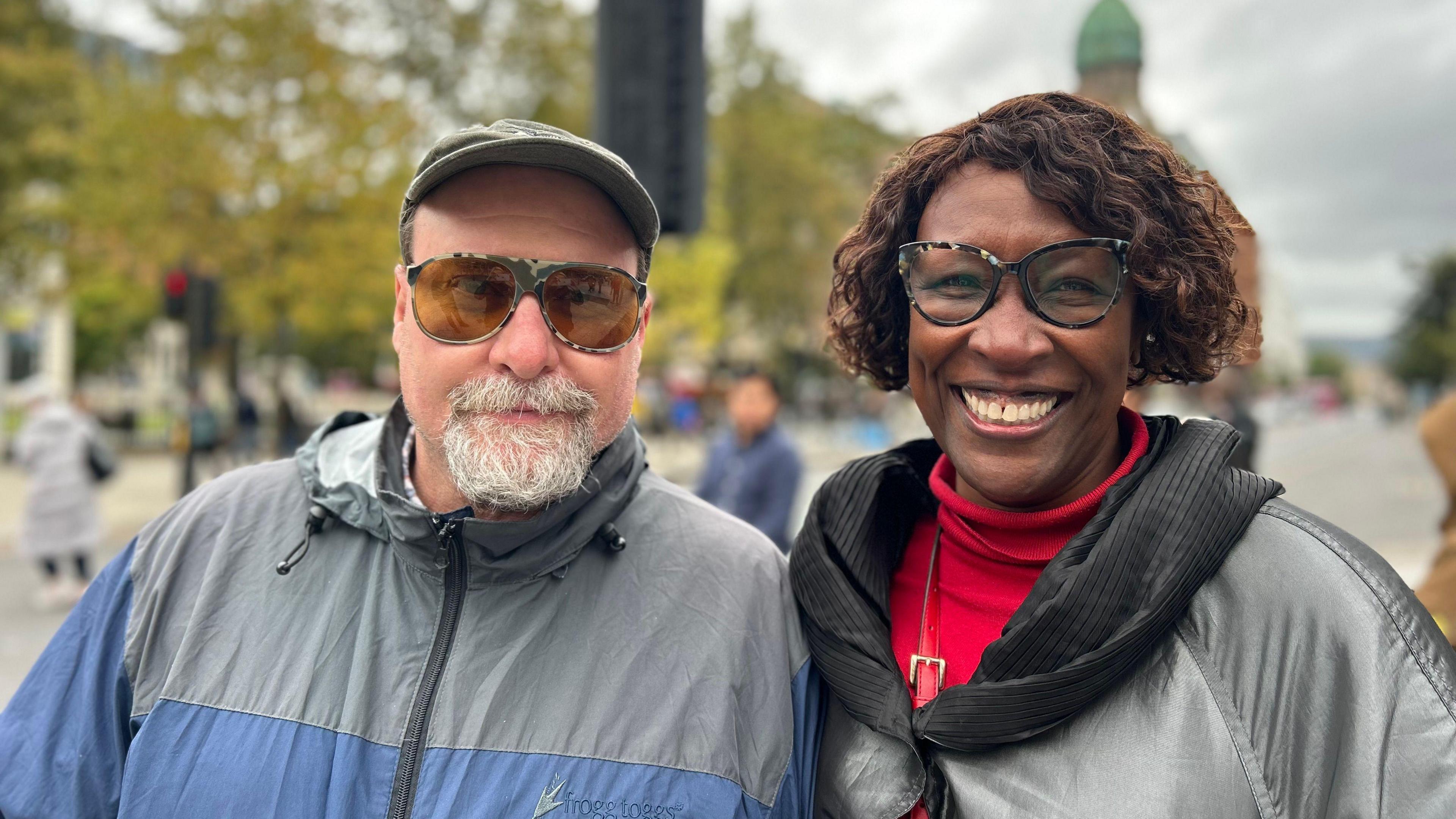 Andy and Alisha Lewis smiling in front of Belfast City Hall. Andy is wearing a raincoat, cap and sunglasses while Alisha is wearing glasses, a red jumper and black coat. 