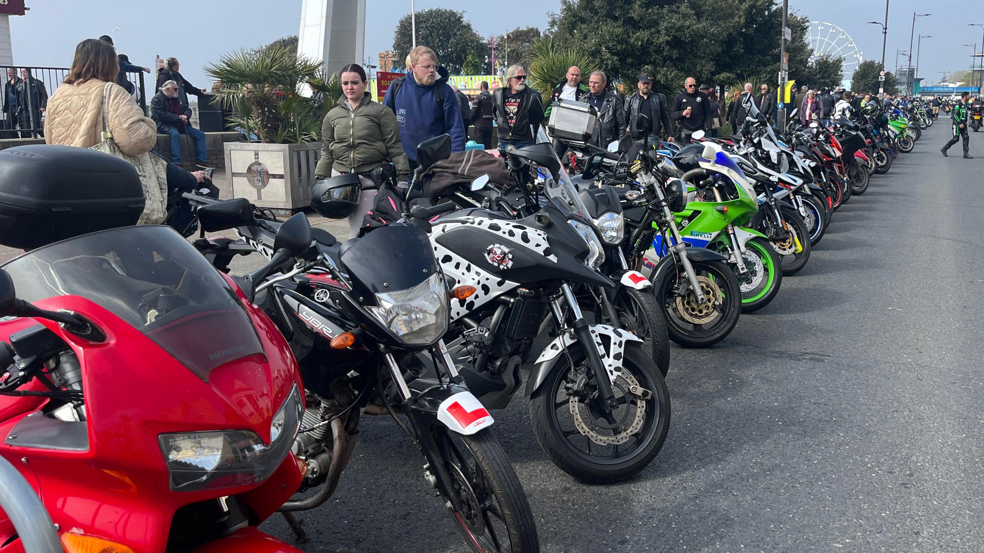 A line of motorbikes parked up on the side of the road. They vary in size and colour. People walk behind them on the pavement. 