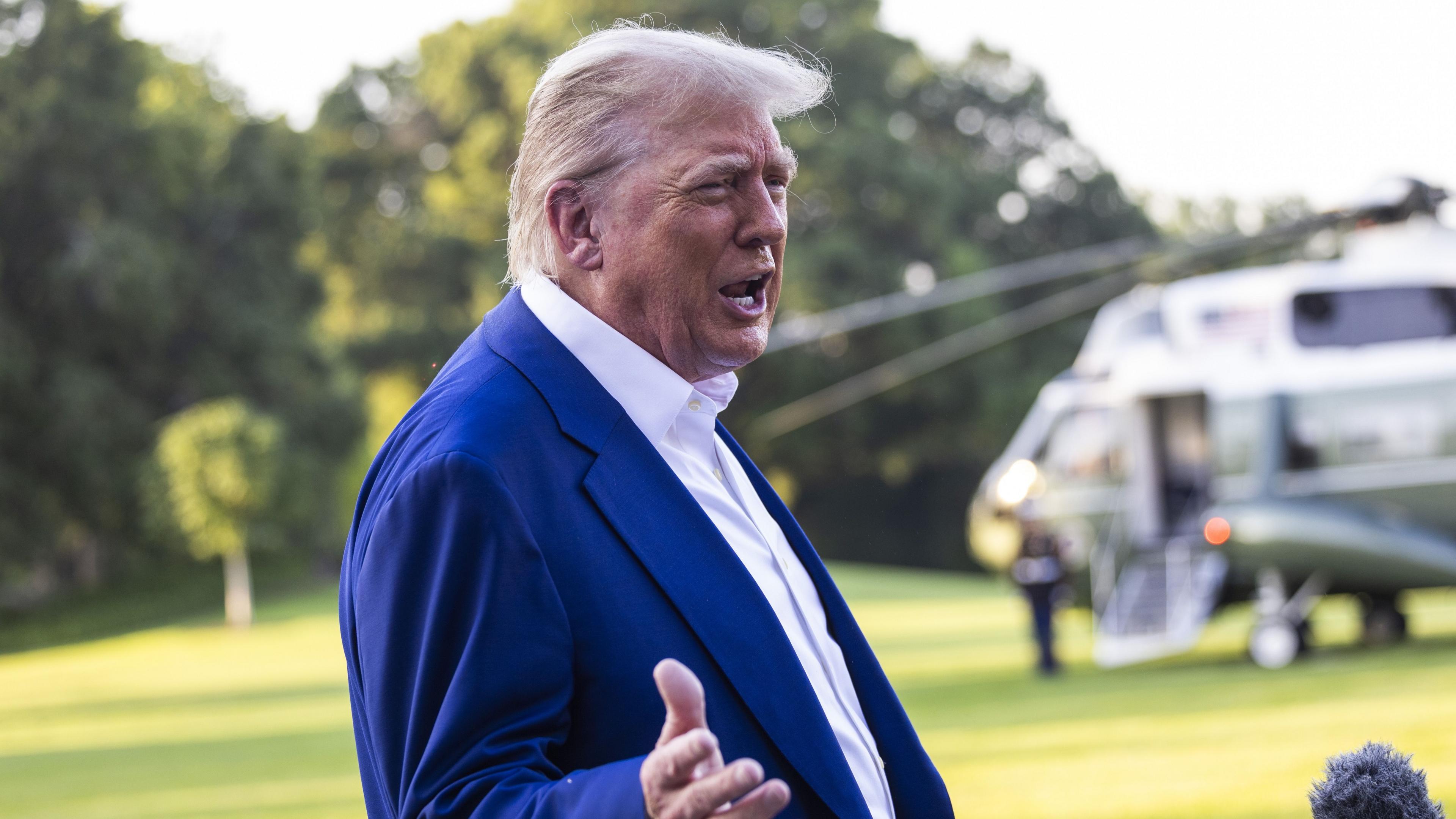 Donald Trump stands in front of a helicopter in the distance and talks. He wears a blue suit and white shirt.