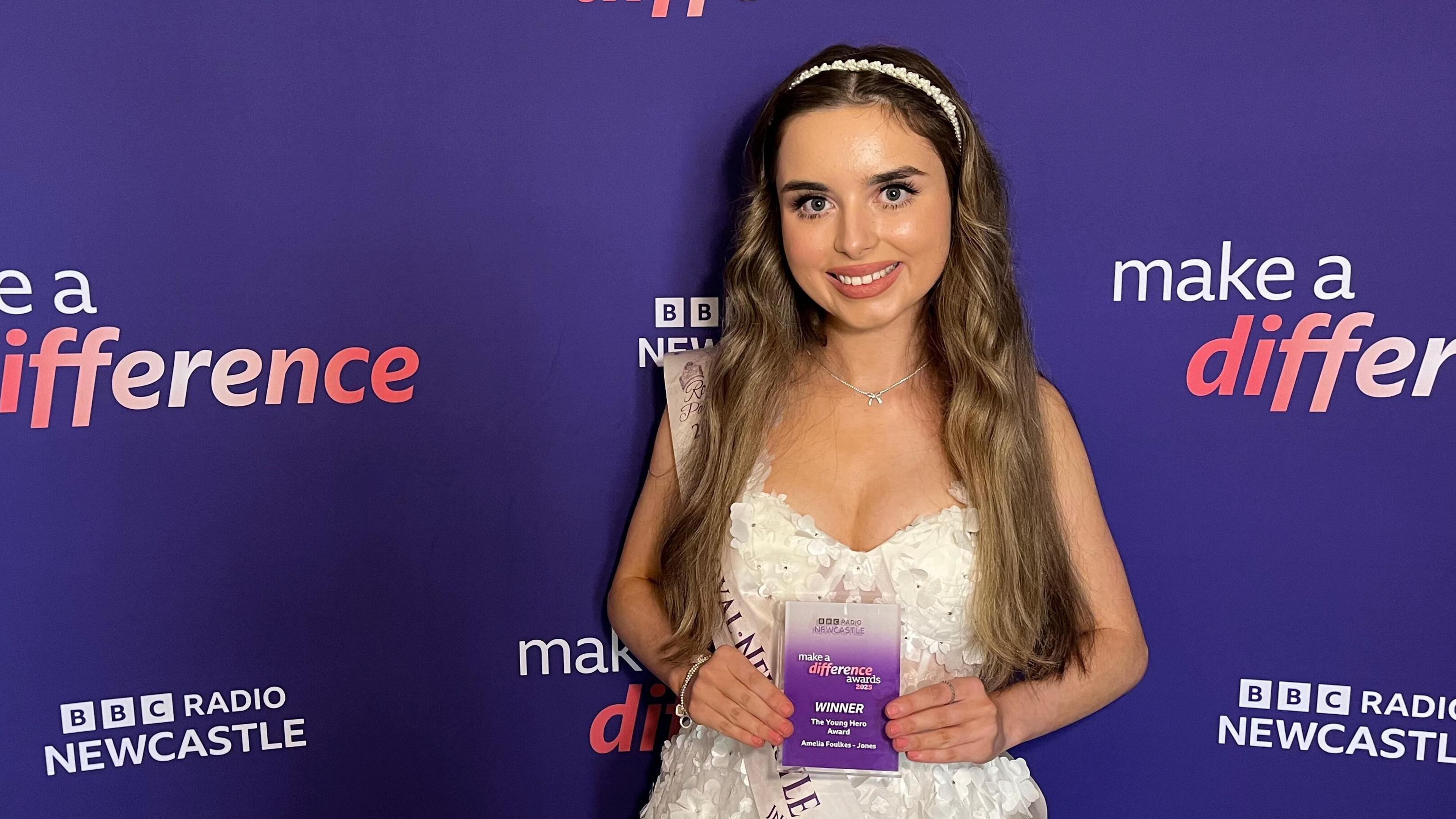 Amelia Foulkes-Jones wears a white evening gown with a pearl headband. She has long brunette hair and is standing with an award in front of purple signage that reads 'MAKE A DIFFERENCE' with the BBC Radio Newcastle branding.
