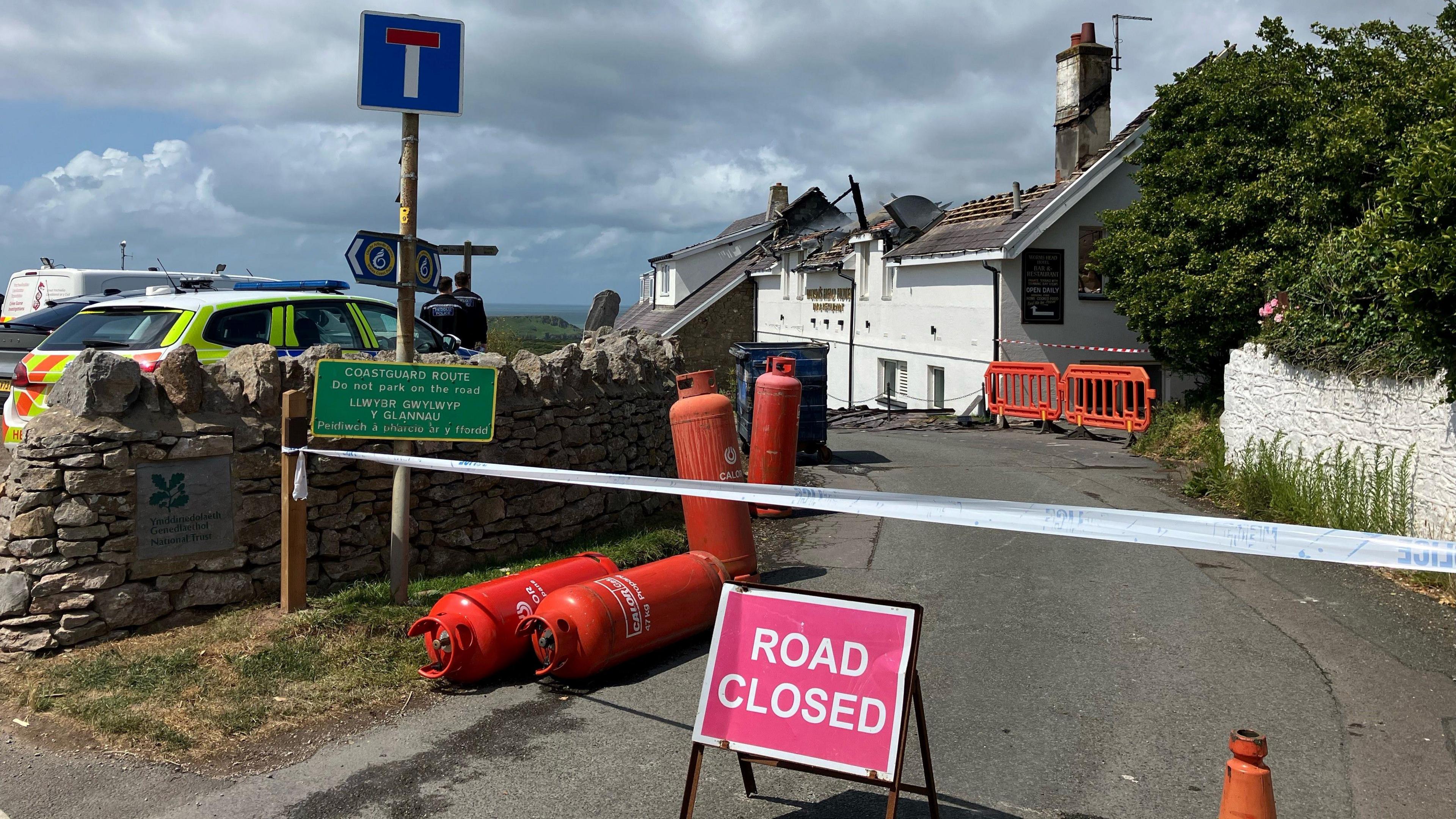 The entrance to the Worm's Head Hotel. There is a road closed sign just at the entrance with police table above it. There is orange canister lying on the ground. The roof of the hotel has been damaged by the fire. There is a police car parked in front of the hotel