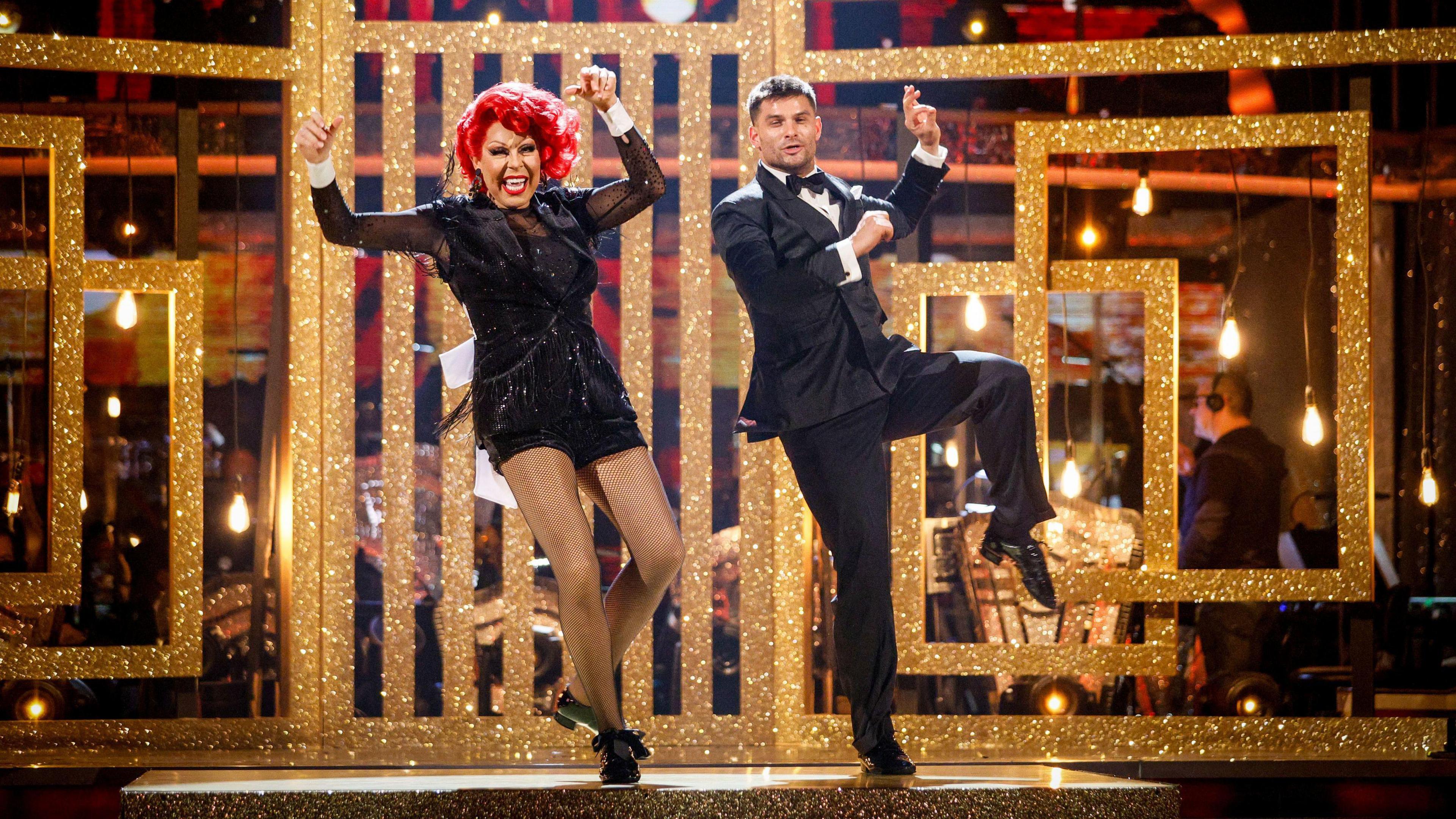 La Voix and Aljaž kick one leg to the side with a golden glittery background behind them