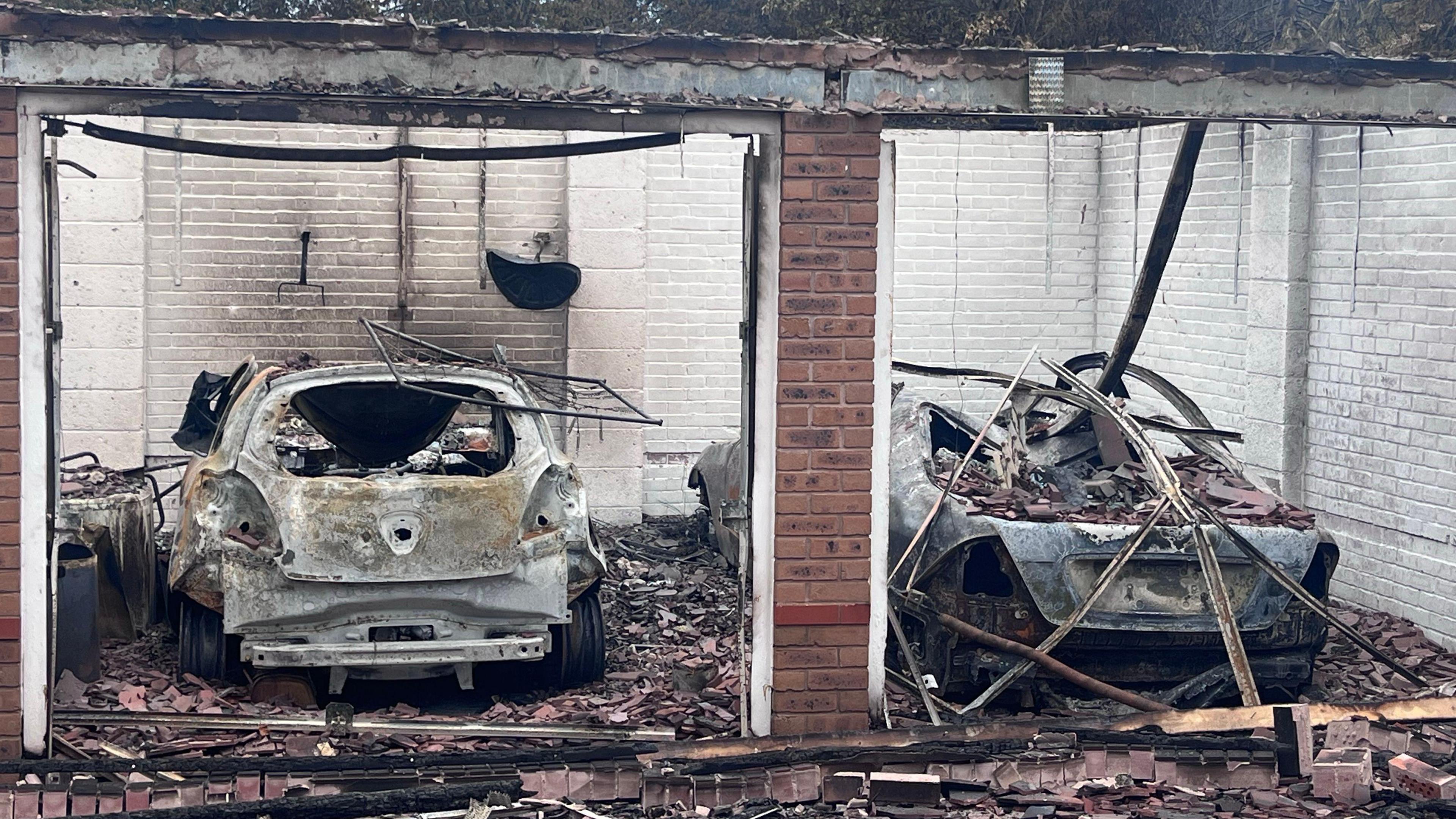 Two cars and a garage which have been completely destroyed by fire. The cars are grey and caved in and there is much material around them. 