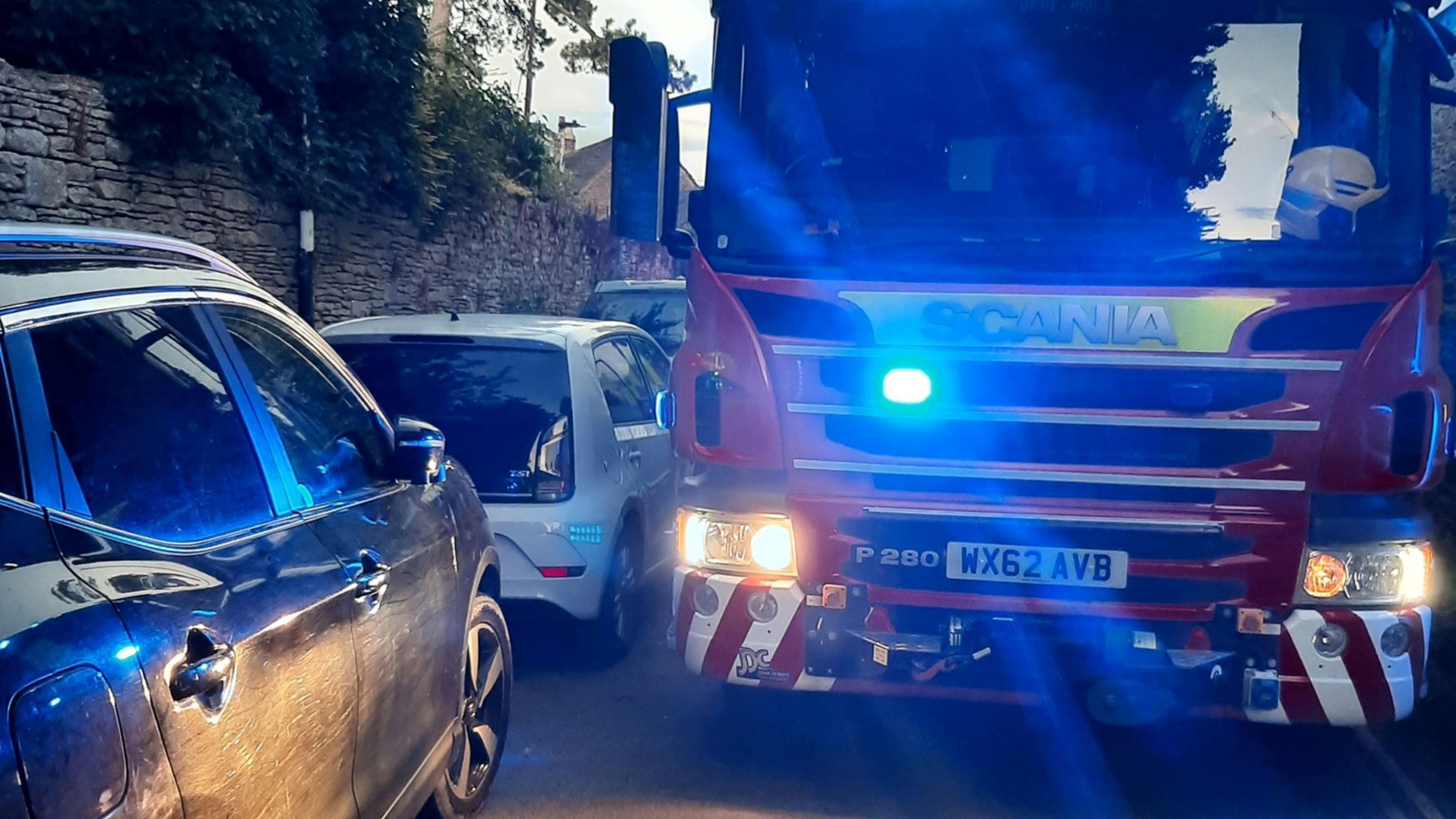 A fire engine with its blue lights navigating down a narrow lane in semi-darkness with vehicles parked against a wall.