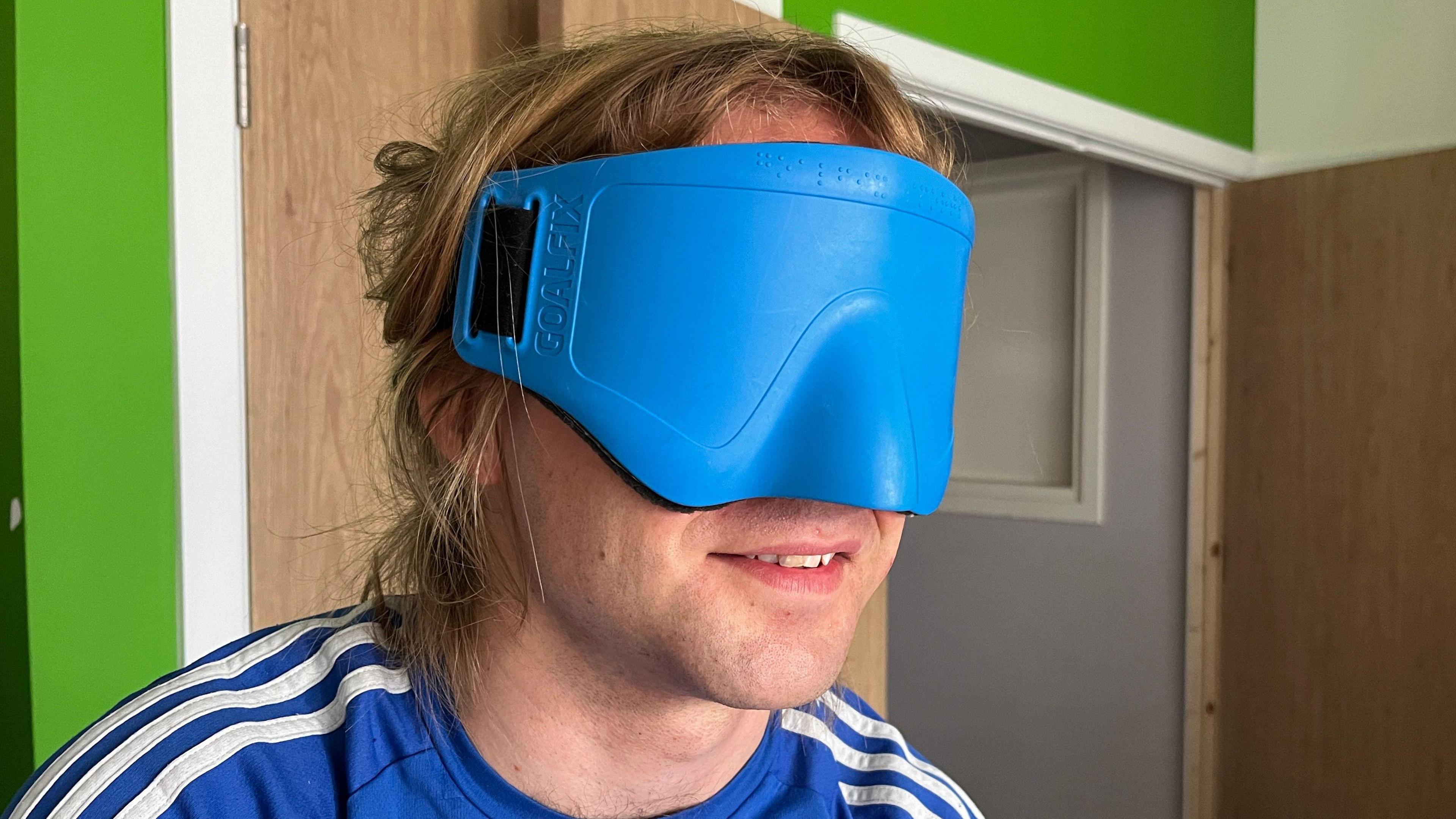 A close up of Warren Wilson's face, as he is wearing a large blue plastic blindfold over his eyes. The middle section of his face is covered, but his mouth is uncovered and he is smiling. He has medium length light blond hair and is wearing a blue top with three white stripes along each shoulder. The room is is in has a bright green wall and wooden door in the corner.