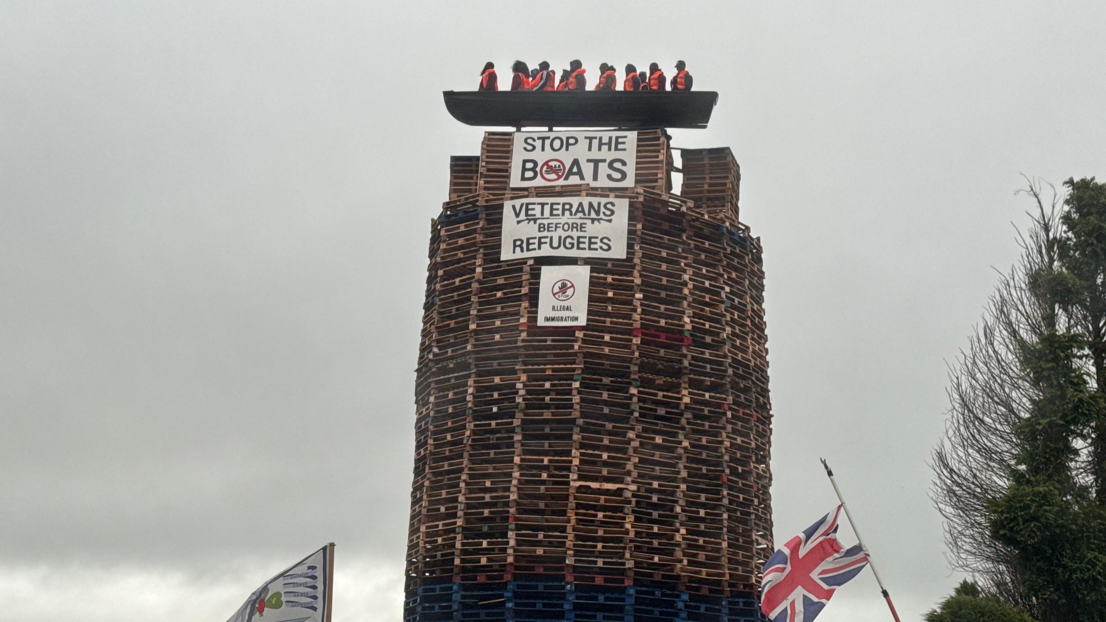 The picture shows a model of a small boat with figures inside on top of a tall bonfire constructed mainly from pallets. Signs saying "Stop The Boats" and "Veterans Before Refugees" are on the bonfire and a Union flag and loyalist flag are at the bottom of the picture