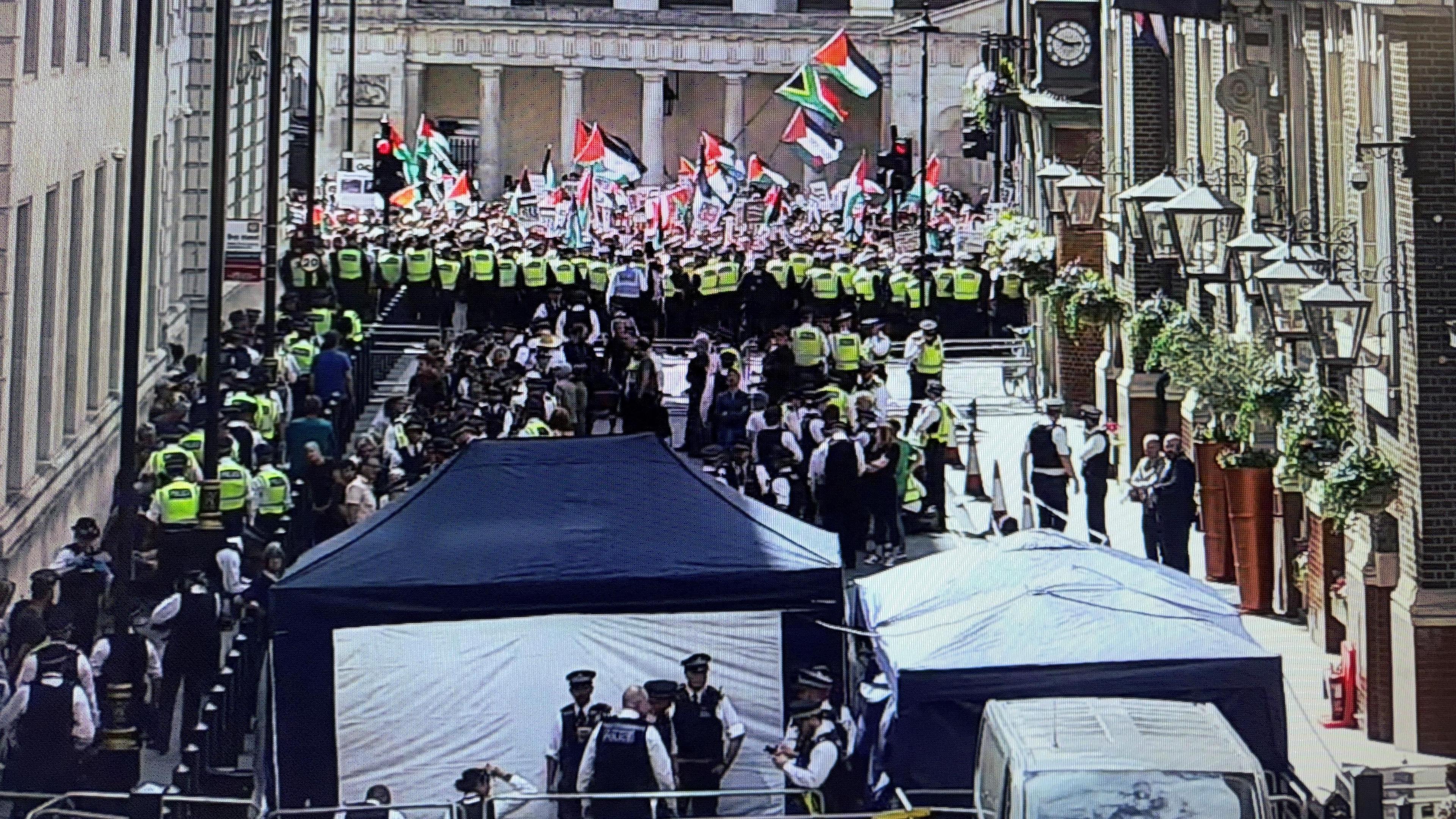 Metropolitan Police arrest processing point in central London at the protest in support of Palestine Action. There are lots of police officers standing near two blue tents. In the background a wall of officers wearing high-vis tops are standing in front of a group of people waving Palestine flags