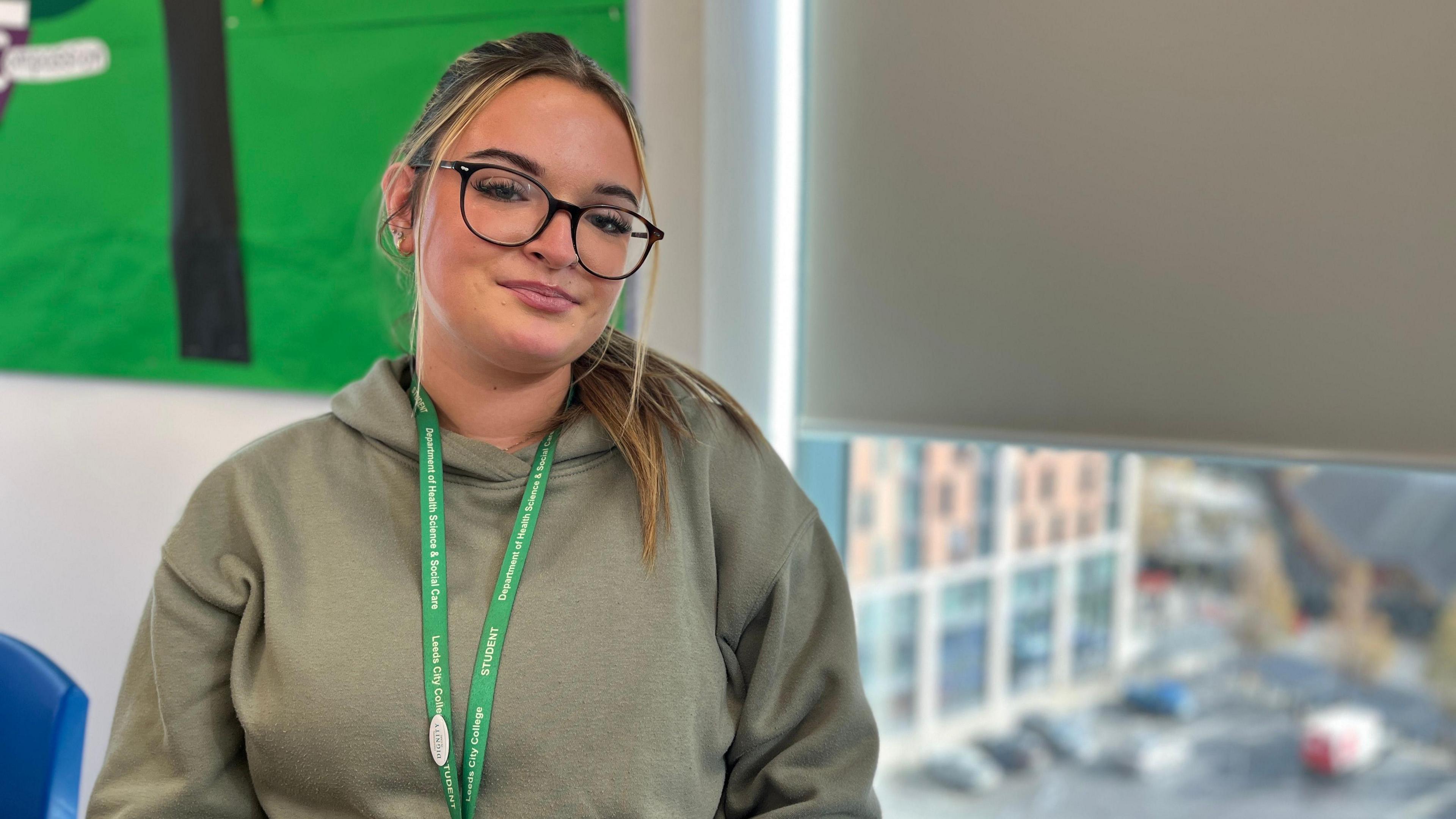 Lola Marshall has long straight blonde hair, which she wears tied back and draped over one shoulder. She wears black glasses and is smiling at the camera. She wears a khaki green hoodie with a greeb Leeds City College lanyard around her neck. She is sat in a room in the college. Behind her is a widow looking out over a carpark, which is out of focus.