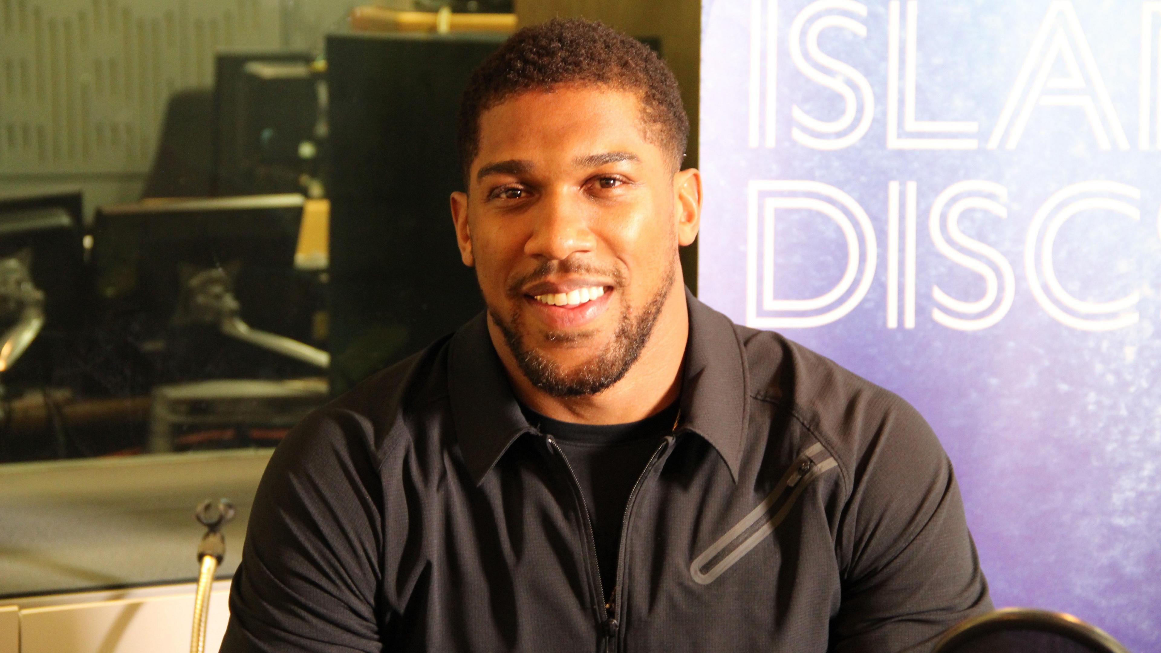 Anthony Joshua smiling at the camera sat in front of a sign saying Desert Island Discs