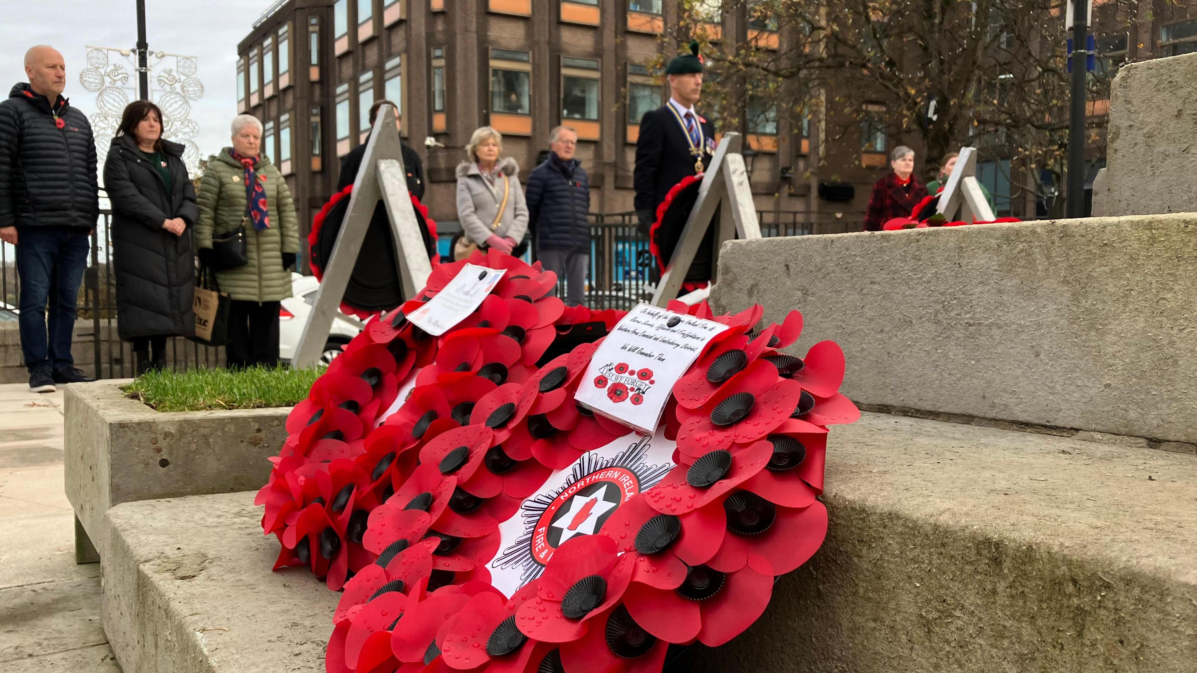 Poppy wreaths laid on the steps of the Cenotaph in Londonderry. In the background several people are gathered observing a moment's silence in remembrance 