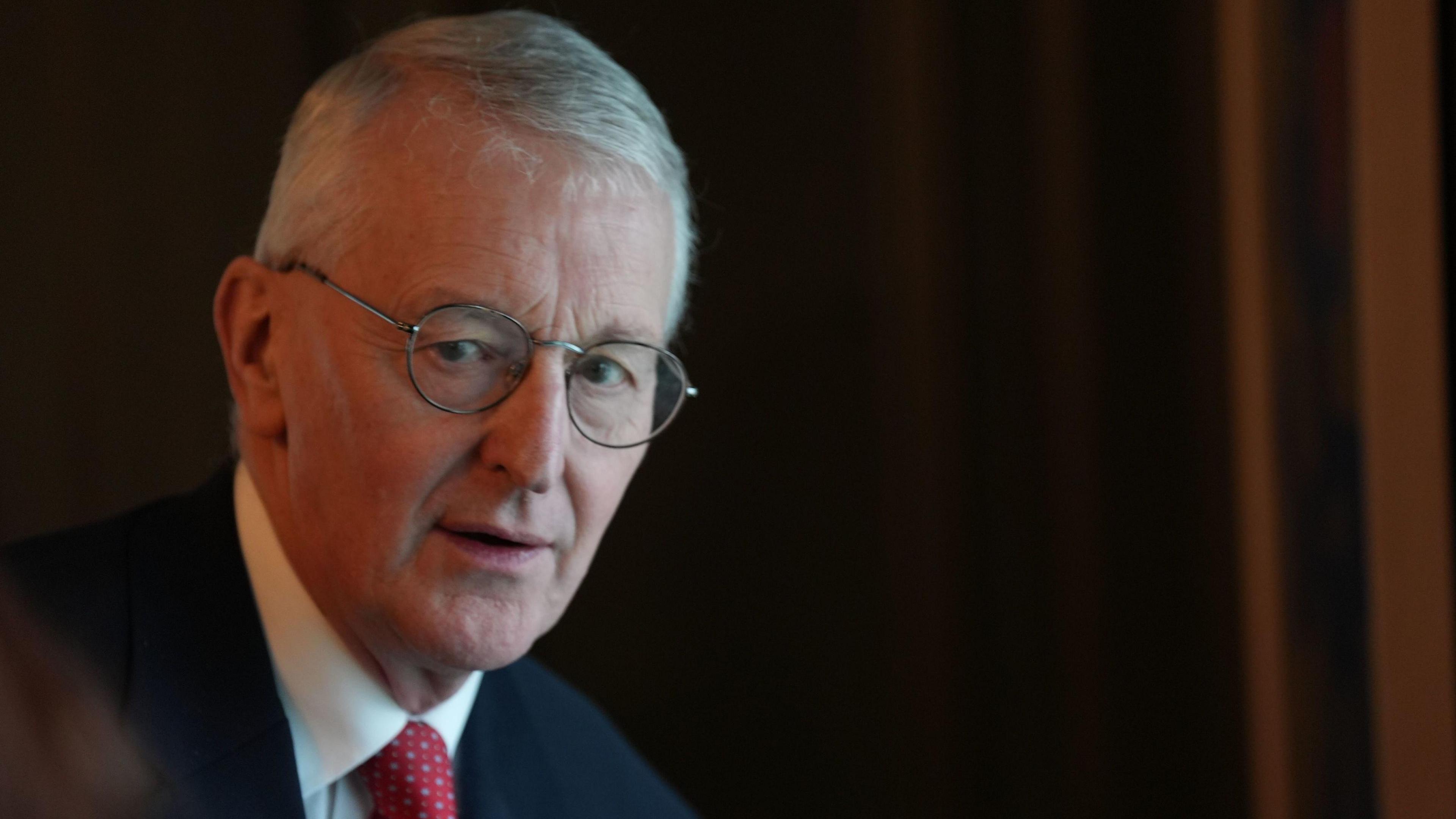 A still image of Hilary Benn as he looks to a person off camera. He has short, greying hair and is wearing a pair of silver, circular glasses. He is wearing a dark suit jacket, a white collared shirt and a red patterned tie.