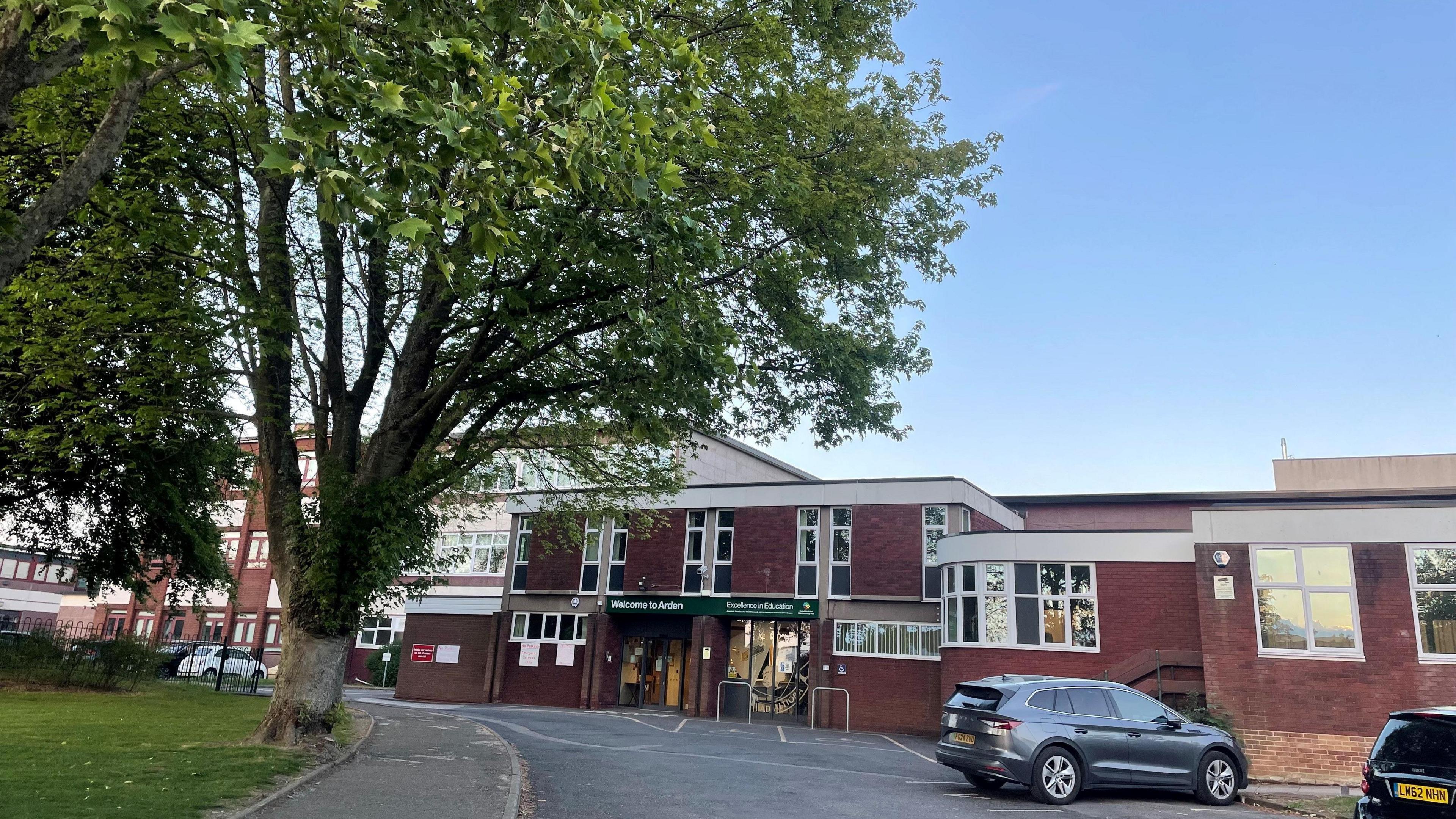 A brick building with dozens of windows. Cars can be seen outside alongside a large green tree.