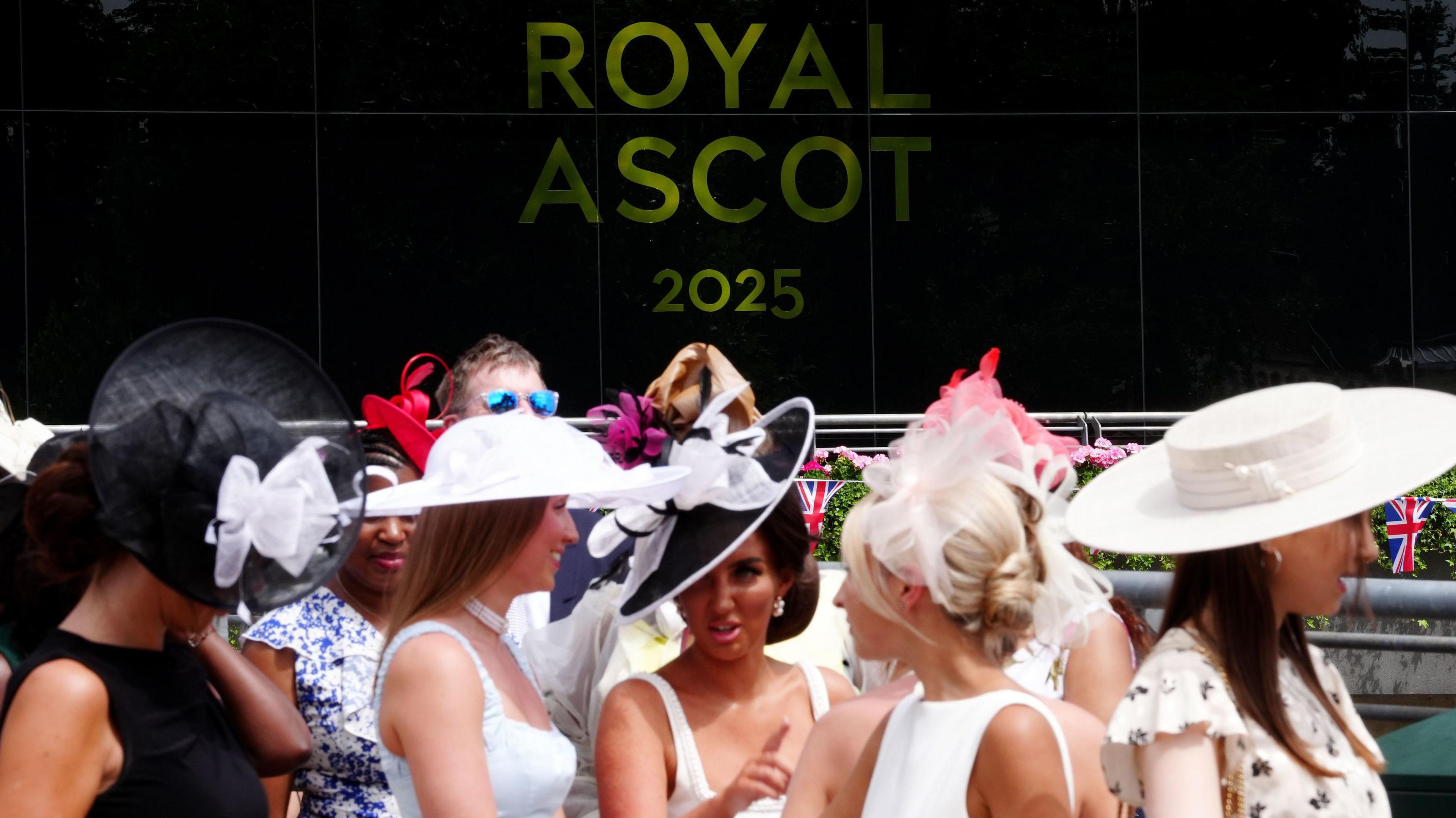 A crowd of women are gathered, all wearing formal dresses and hats talking to one another. In the background is a large black sign with yellow writing on it which reads Royal Ascot 2025.