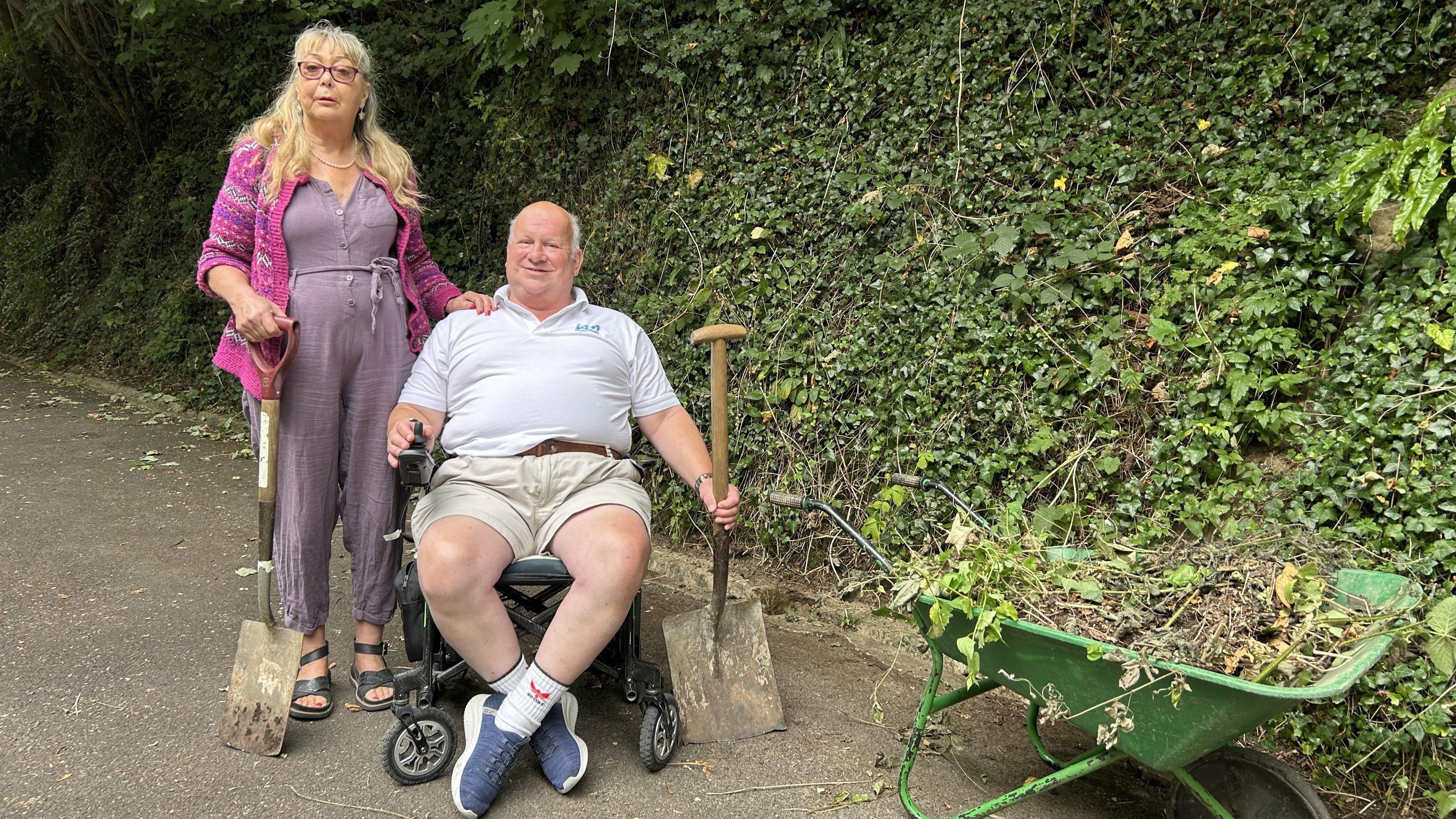 Peter Davis, who is wearing shorts and T-shirt and is sitting in a wheelchair, with Monica standing close by. She is wearing a purple bodysuit and is holding a spade. They are both near a wheelbarrow with leaves and twigs inside.