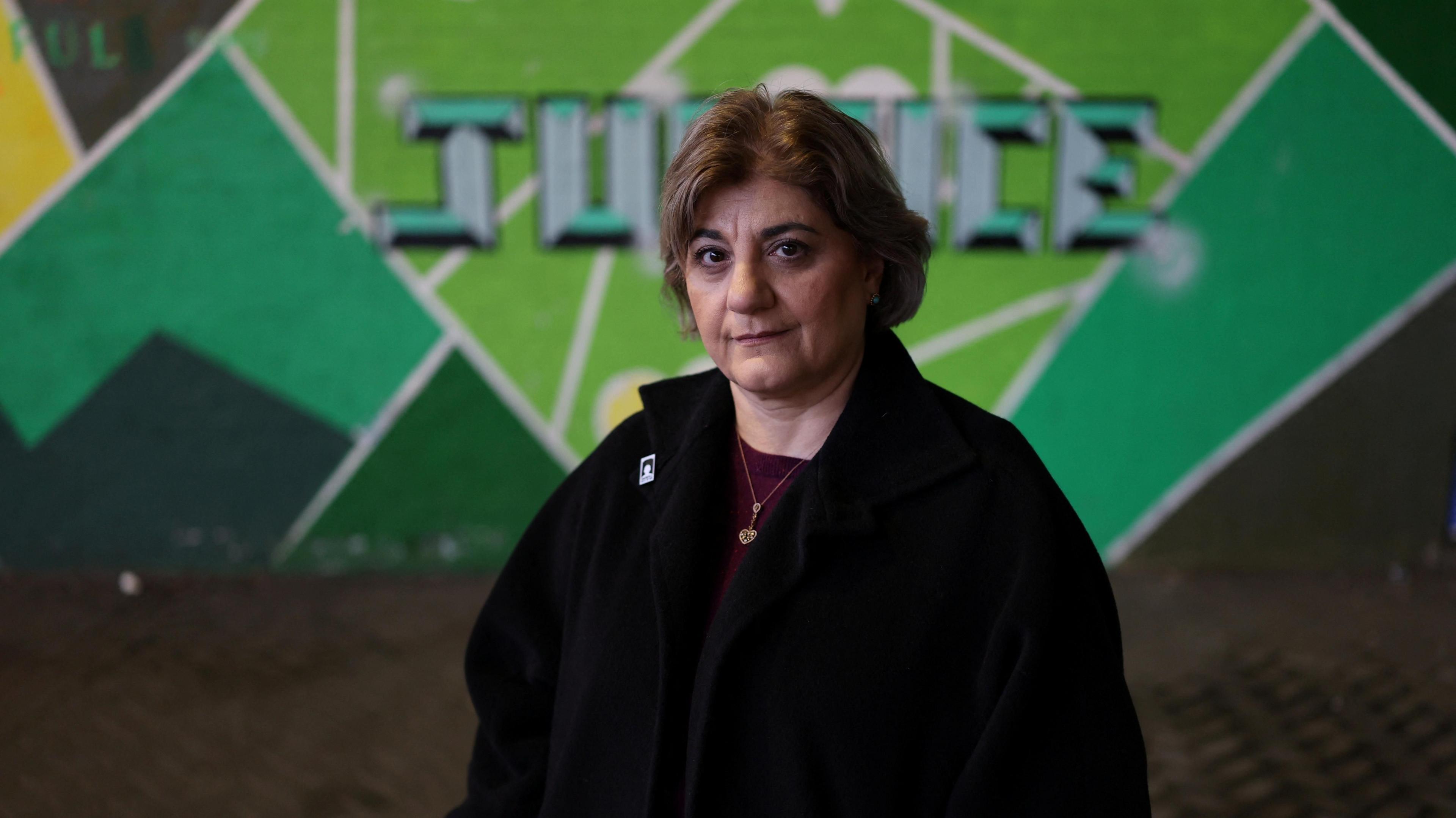 Kimia Zabihyan, a woman with short brown hair wearing a black fleece and maroon top, stands in front of a green mural that says 'justice'.