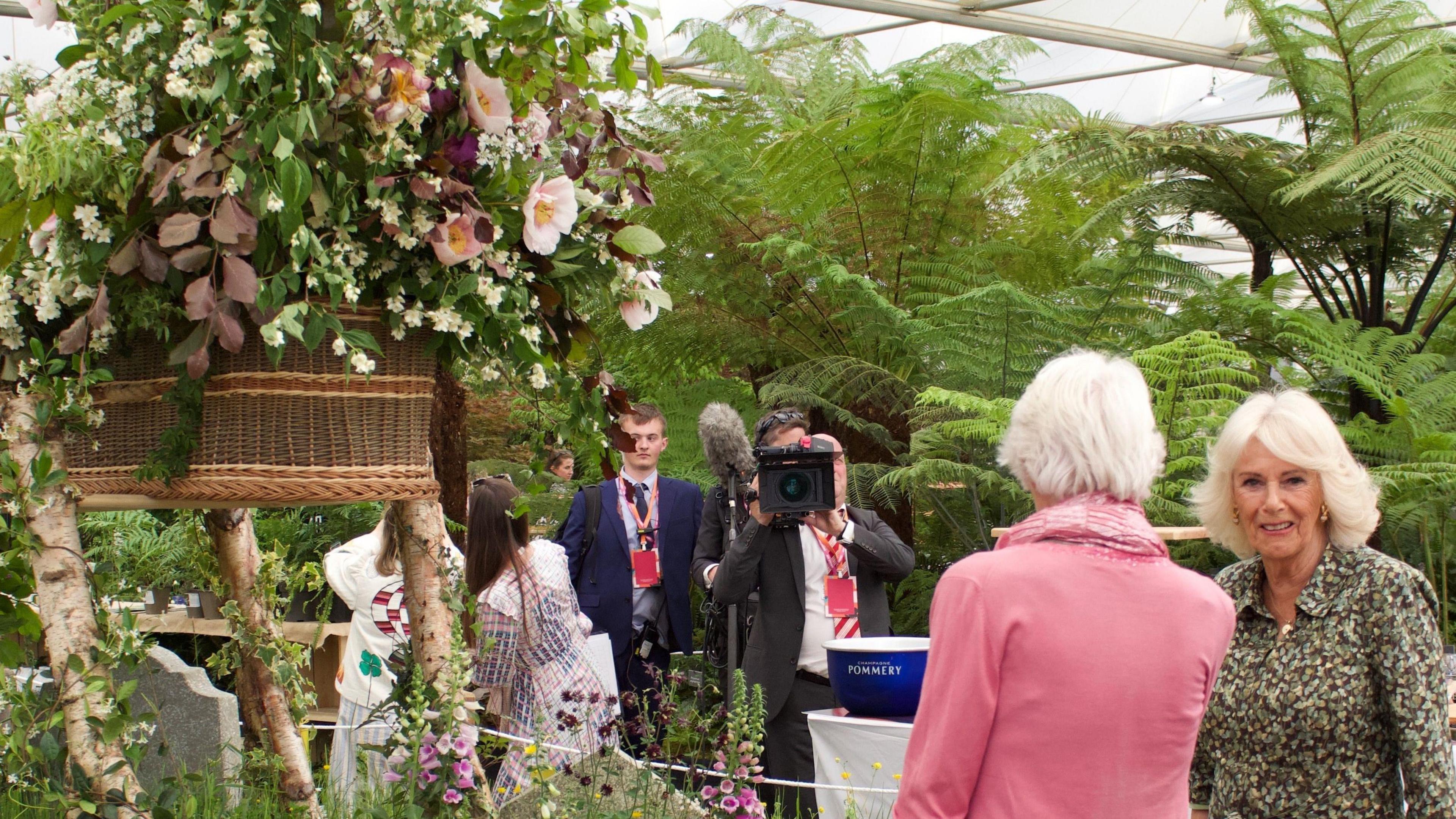 The coffin of flowers in the above picture, from a different angle. Queen Camilla, a woman with shoulder-length white hair and wearing a patterned blouse, stands nearby, next to a woman with short white hair wearing a pink jacket and whose back is to the camera. Other people are in the background, one of whom is holding a video camera.